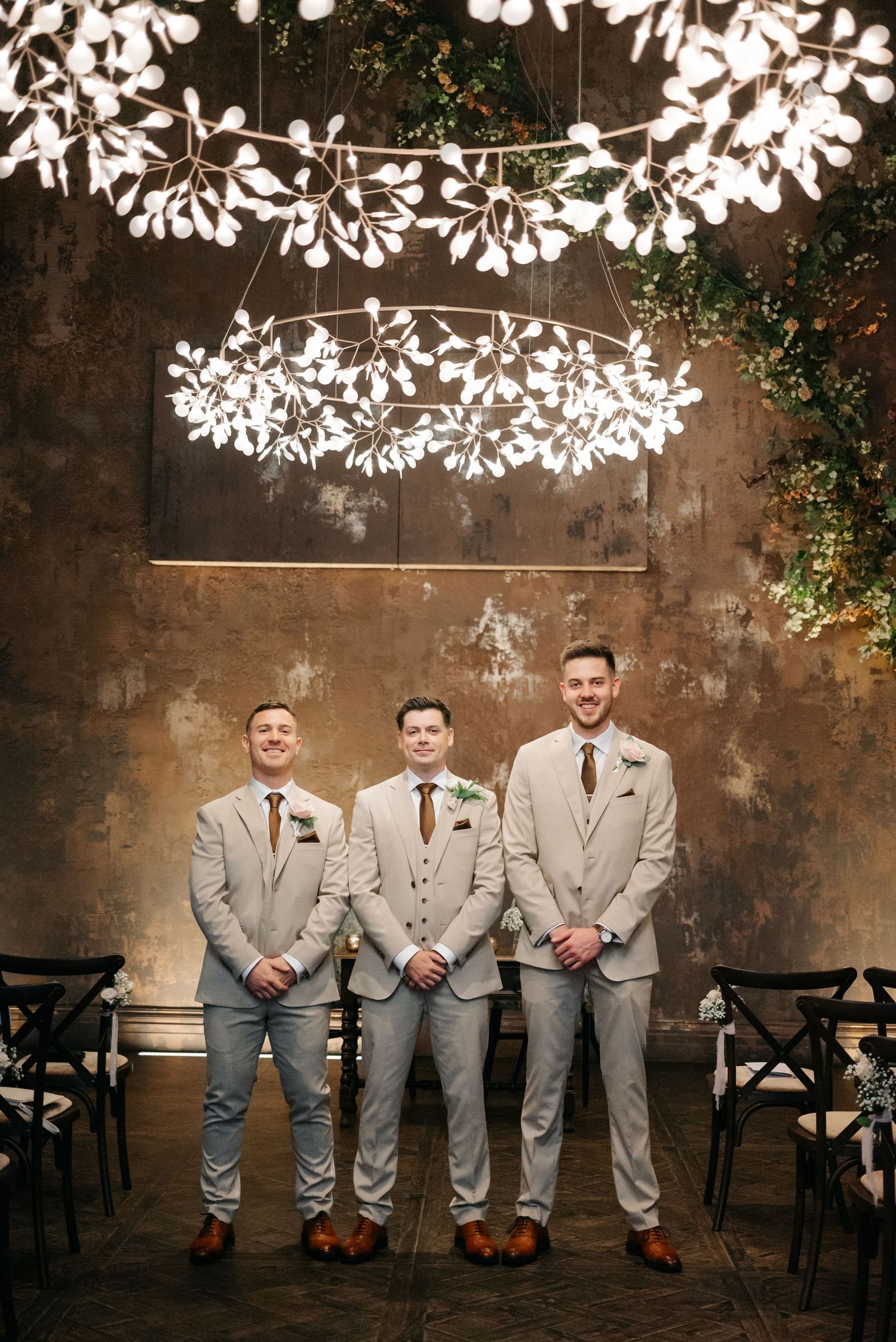Groomsmen in matching light gray suits pose beneath decorative white floral ceiling installation.