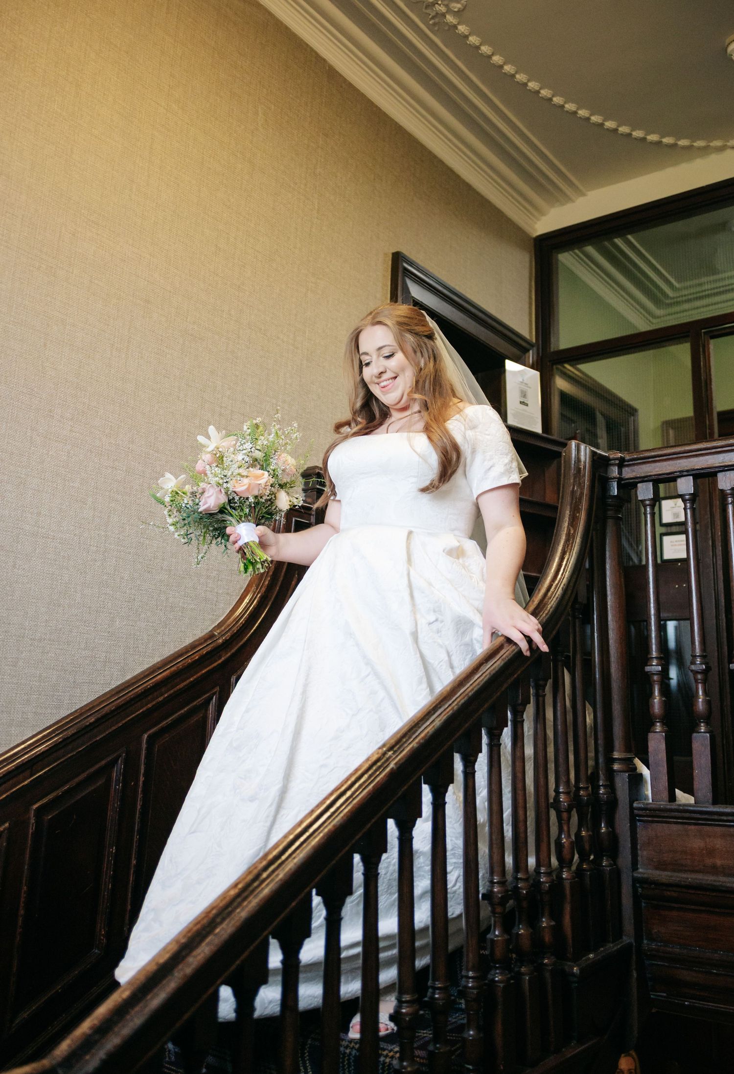 A bride in a white wedding gown poses elegantly on a wooden staircase with a floral bouquet.