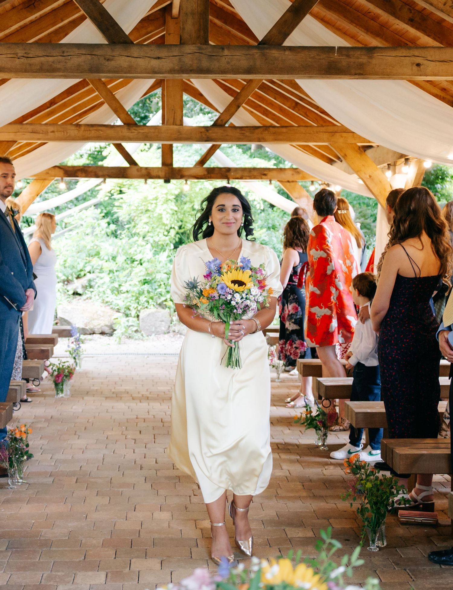Wedding guests gather under a rustic wooden pavilion decorated with floral arrangements.