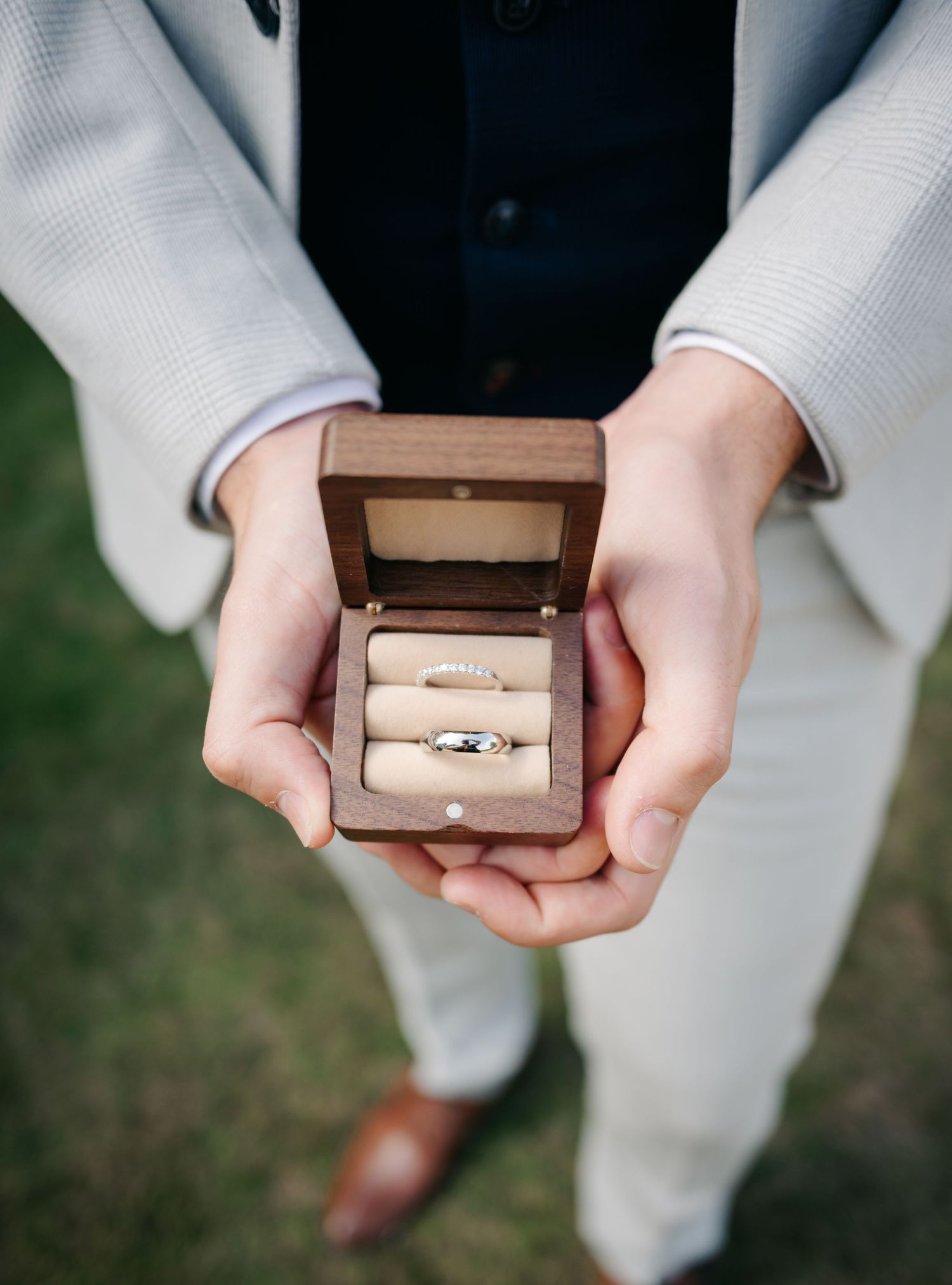 Hands holding a brown wooden ring box containing two wedding bands against a light grey background.
