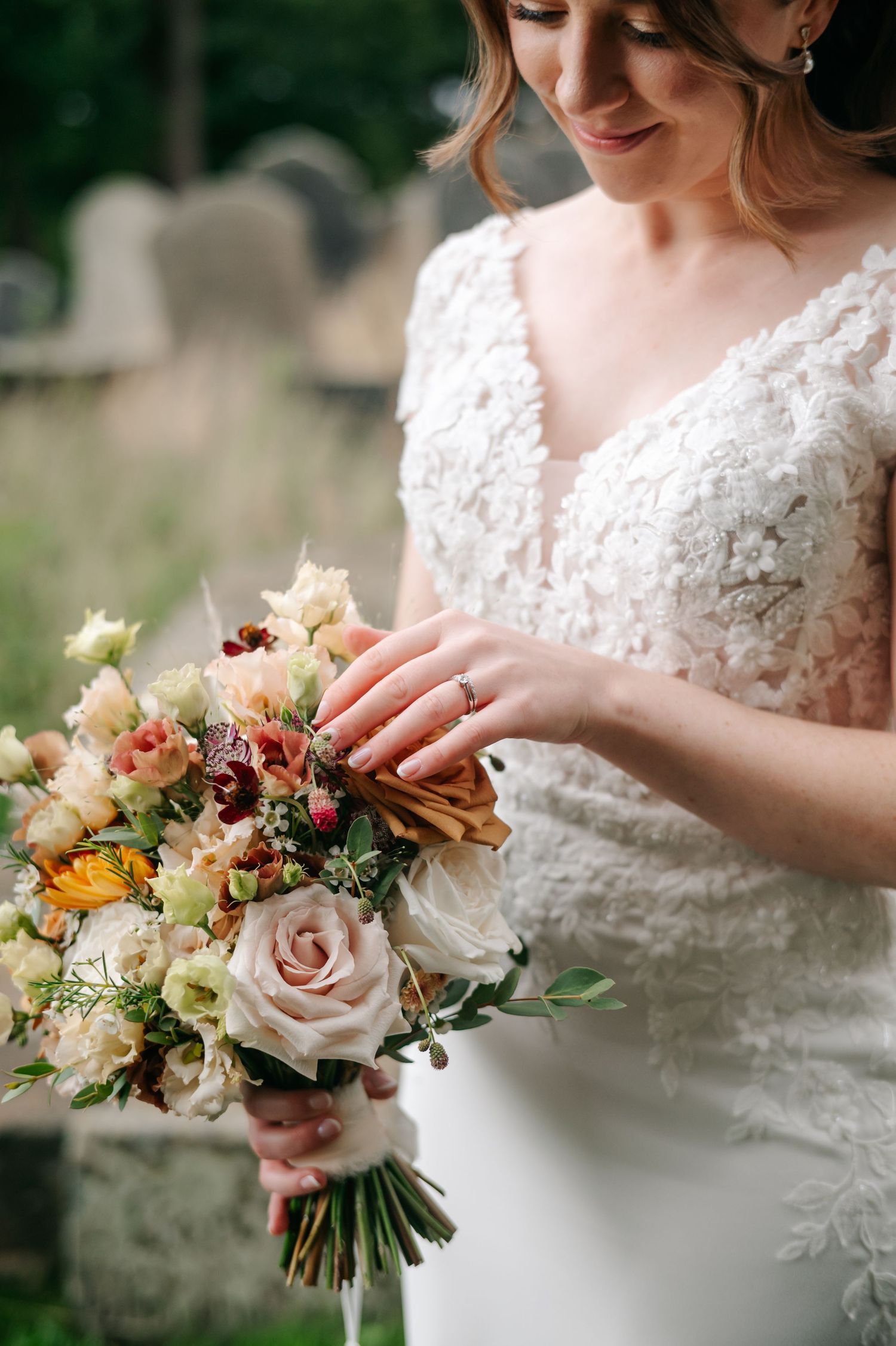 Bride in a lace V-neck wedding gown holds a romantic bouquet of blush roses and mixed garden flowers, with an engagement ring visible.