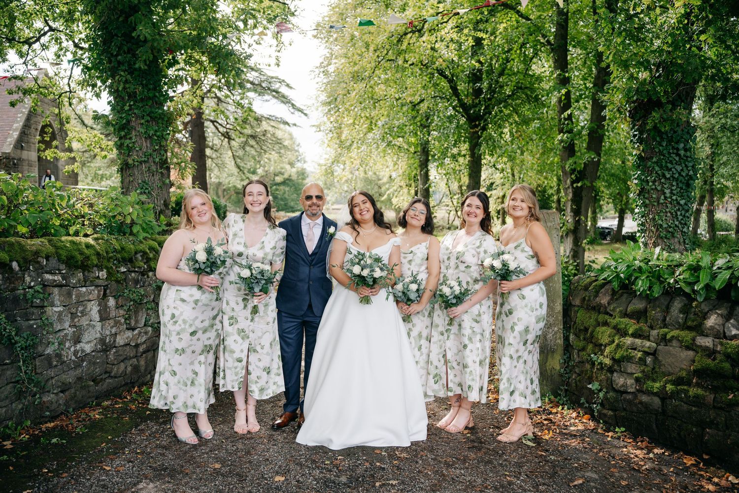 Wedding party poses outdoors in floral bridesmaid dresses and formal attire against a lush green garden backdrop.