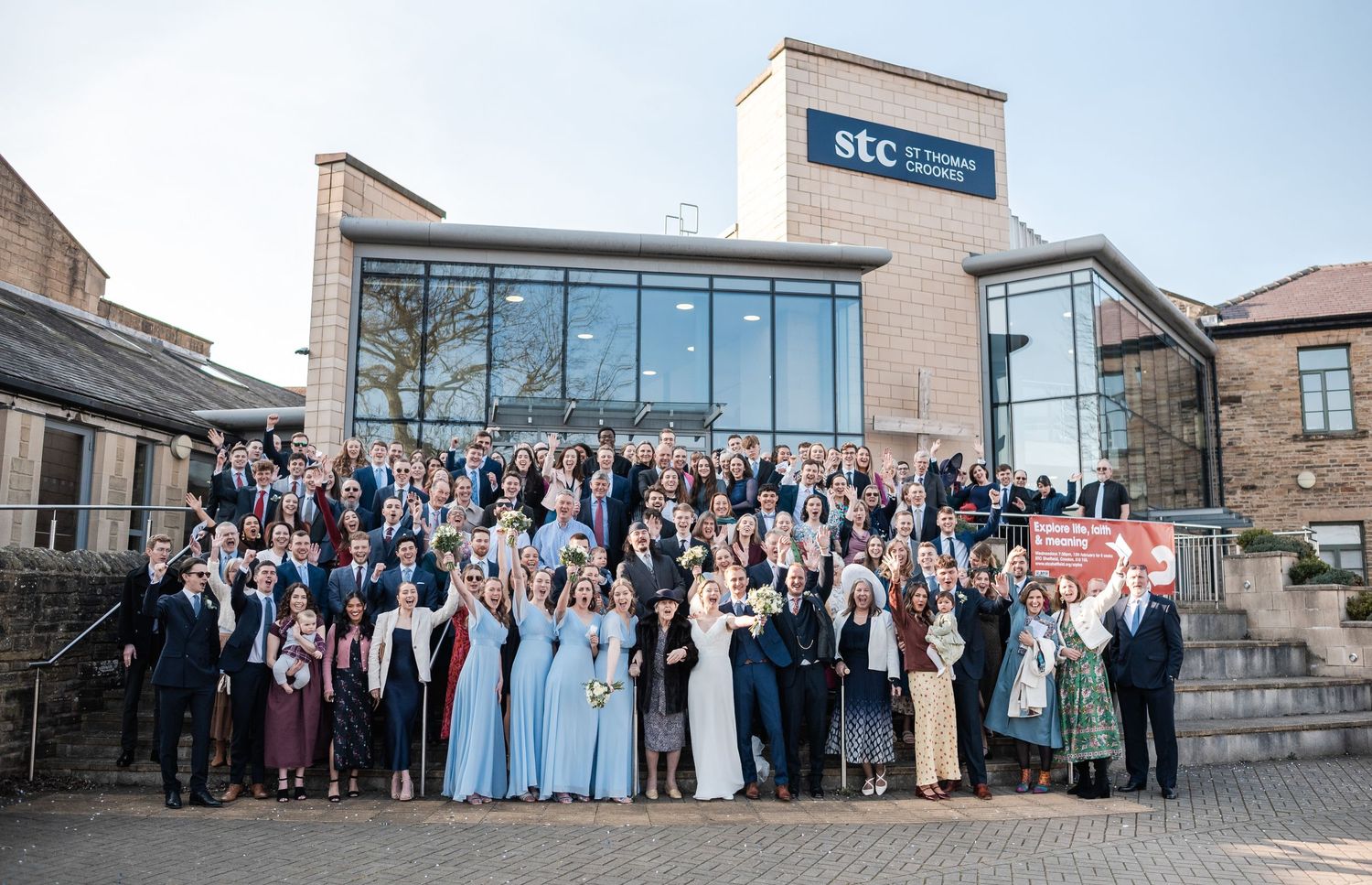 Large wedding party group photo taken outside modern brick building with glass facade.