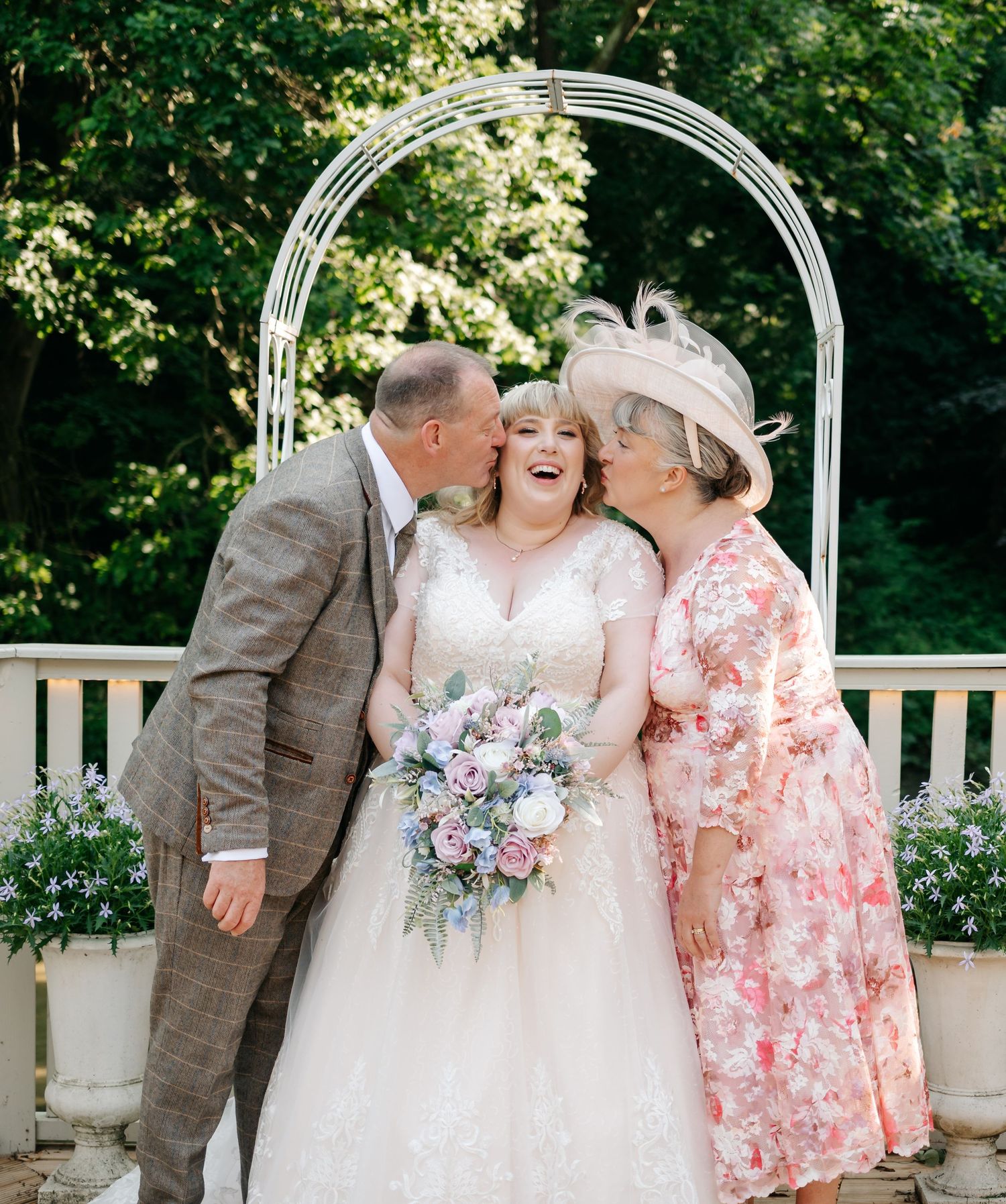 Bride laughing joyfully as both parents kiss her on the cheeks under a white arch outdoors.