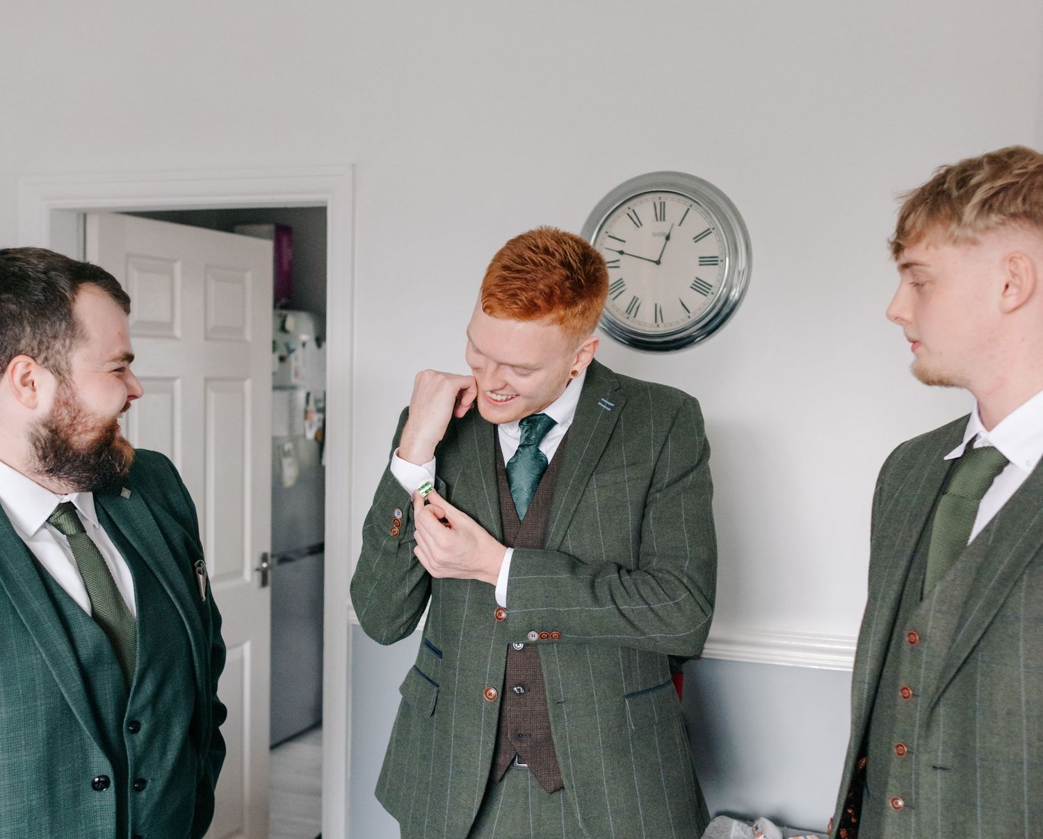 Three men in matching green suits stand together in a room with a white wall and clock.
