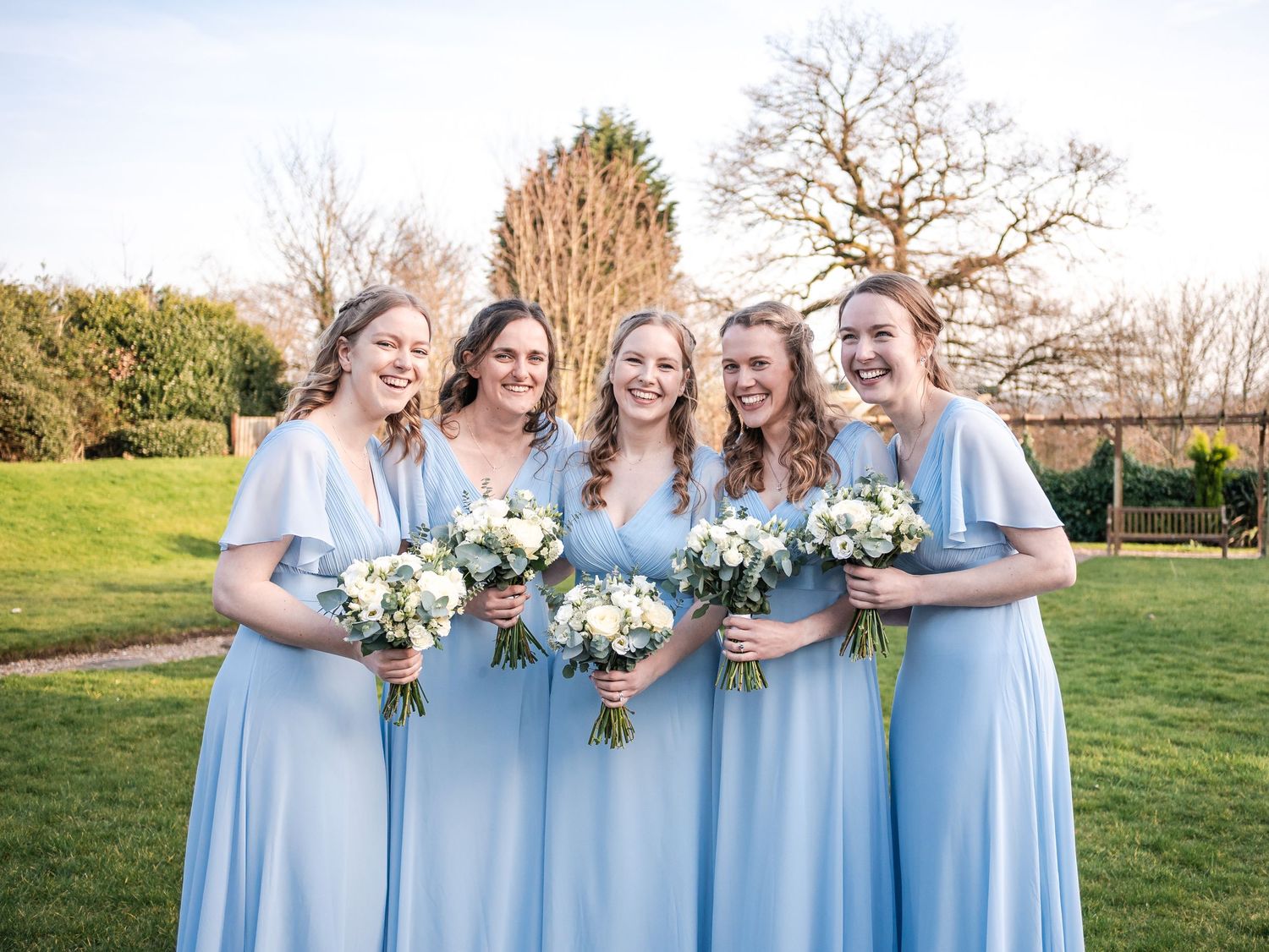 A group in matching light blue bridesmaid dresses hold white floral bouquets while standing together outdoors.
