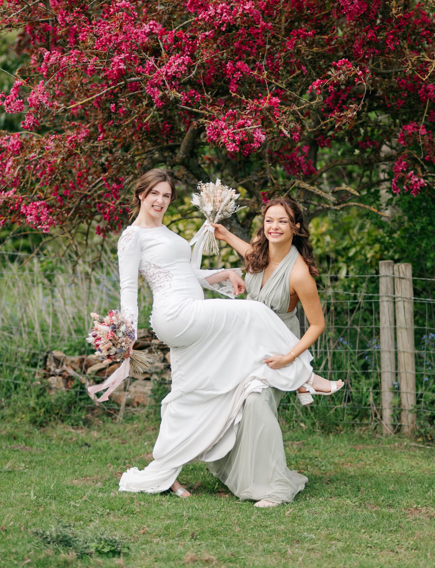 A couple in white dresses dances playfully on grass beneath vibrant pink flowering trees in a garden setting.