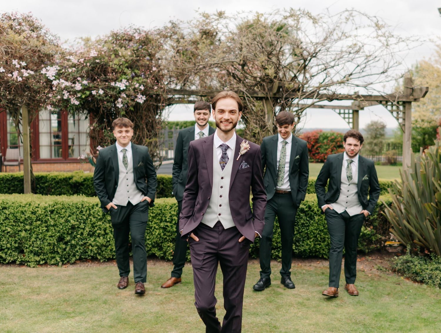 Groomsmen in formal attire pose playfully in a garden setting during a wedding celebration.