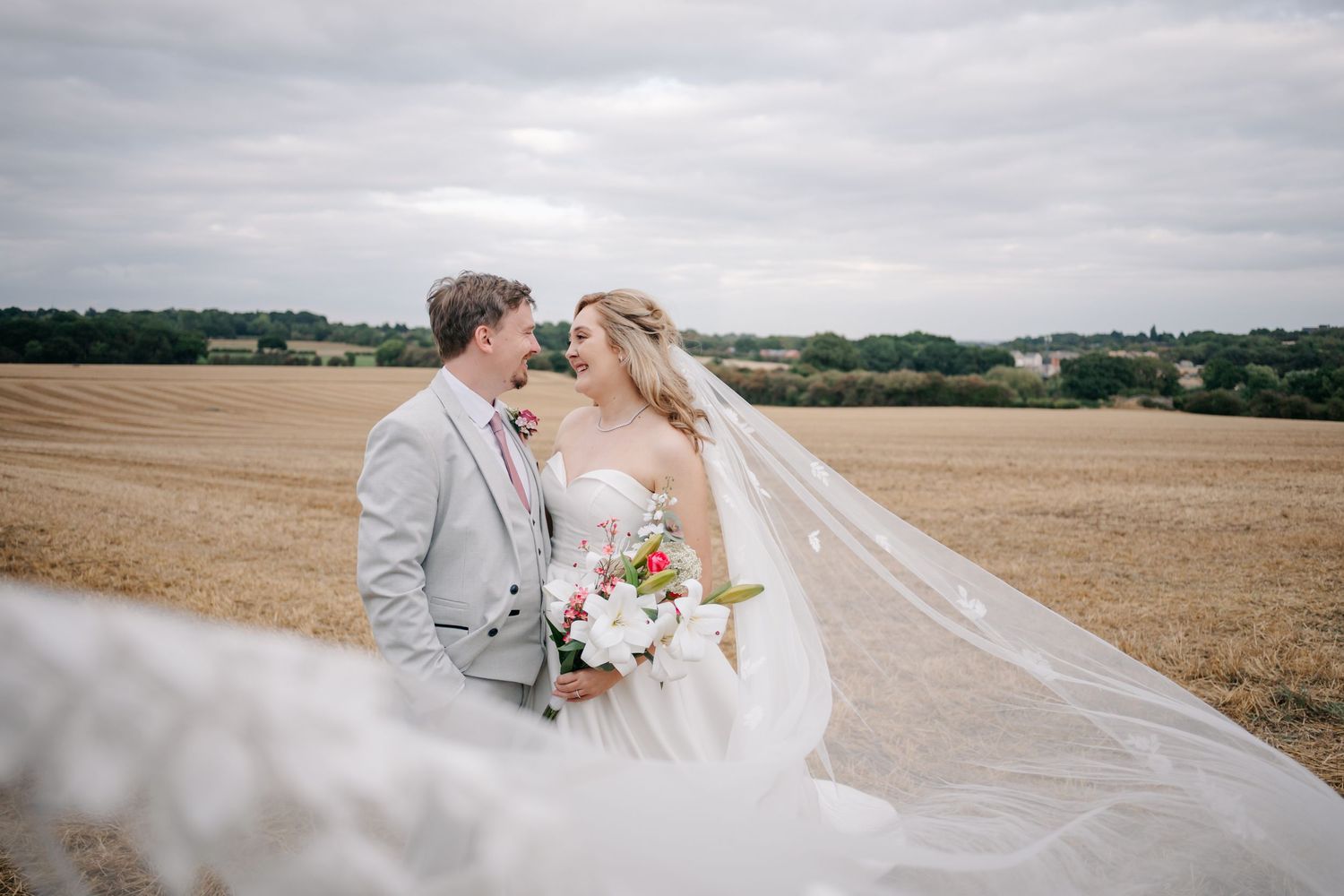A couple in wedding attire embraces in a rustic wheat field on an overcast day.