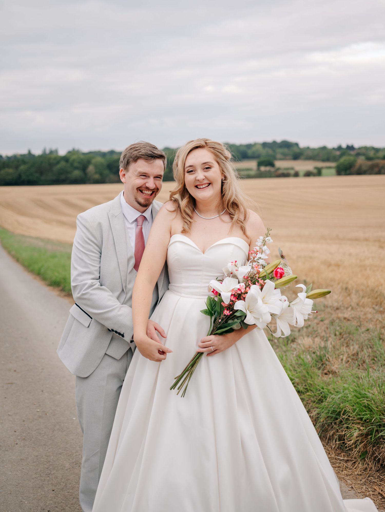 Happy newlywed couple posing on a rural road, bride in white gown holding bouquet, groom in gray suit.