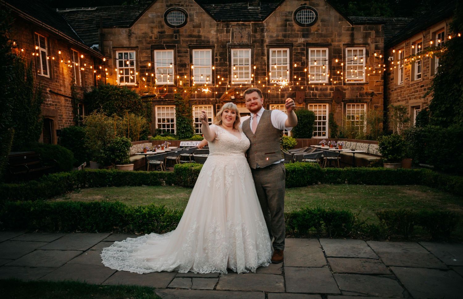 Bride and groom posing at dusk outside stone venue with fairy lights and garden seating.