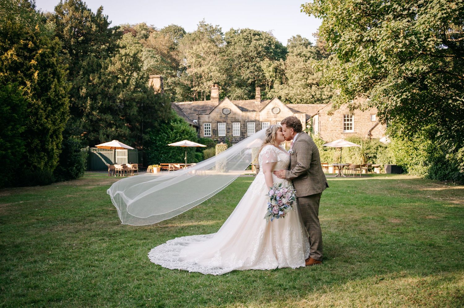 A bride and groom share a romantic kiss on a lawn, her veil flowing in the breeze.