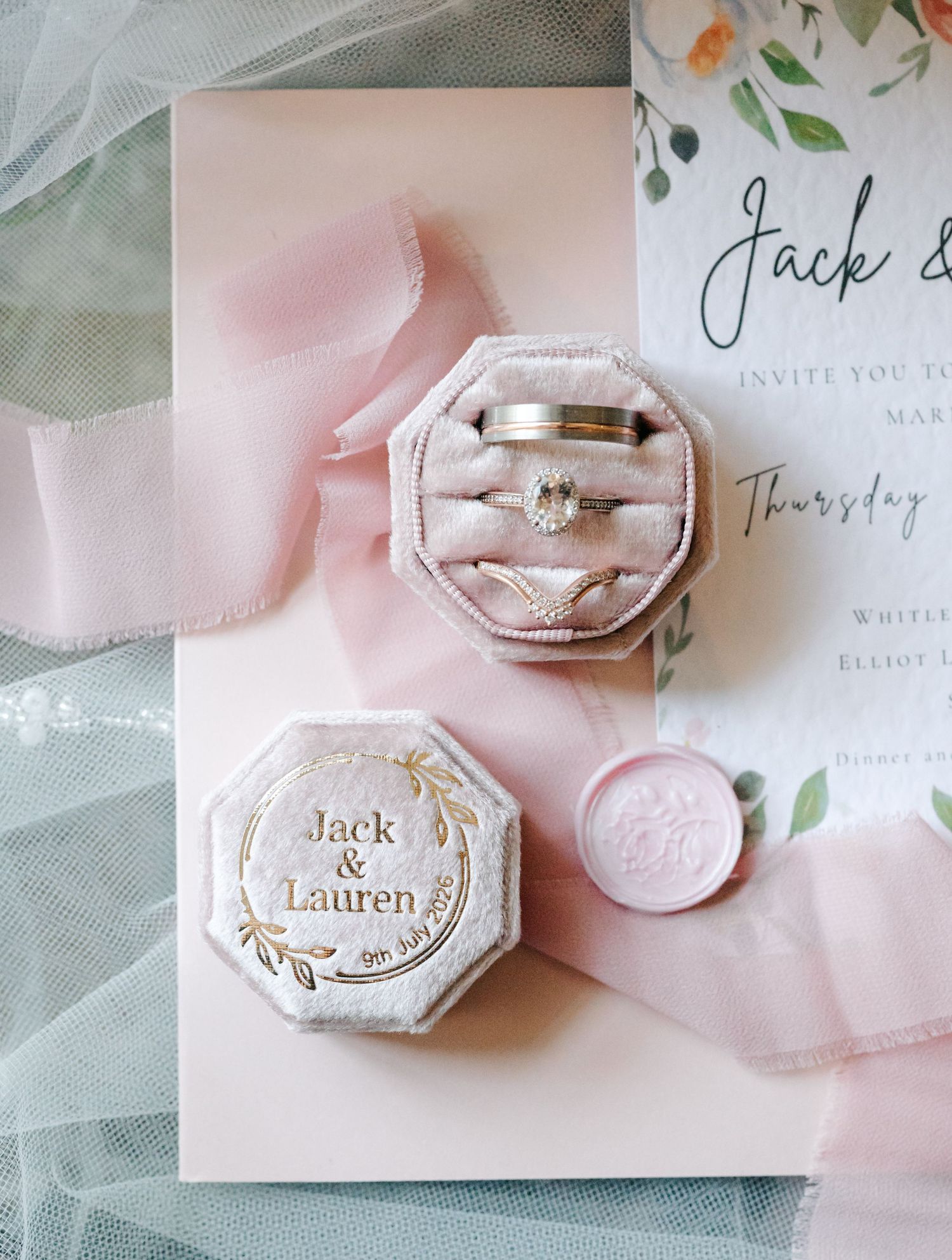 Elegant wedding flat lay with velvet ring box engraved Jack & Lauren, engagement rings, pink invitation, and silk ribbon.