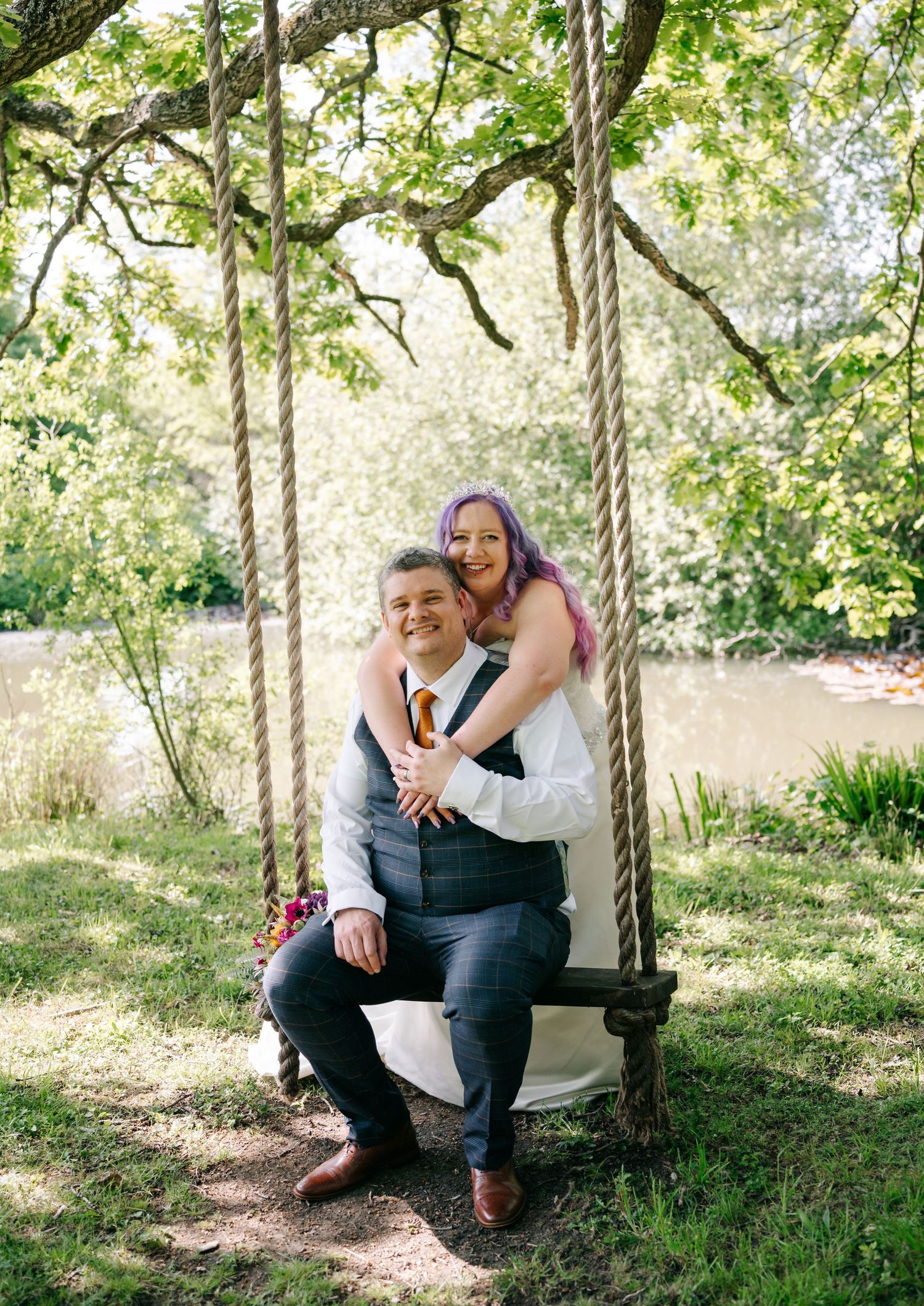 A bride and groom pose on a wooden rope swing surrounded by lush greenery near a pond on their wedding day.