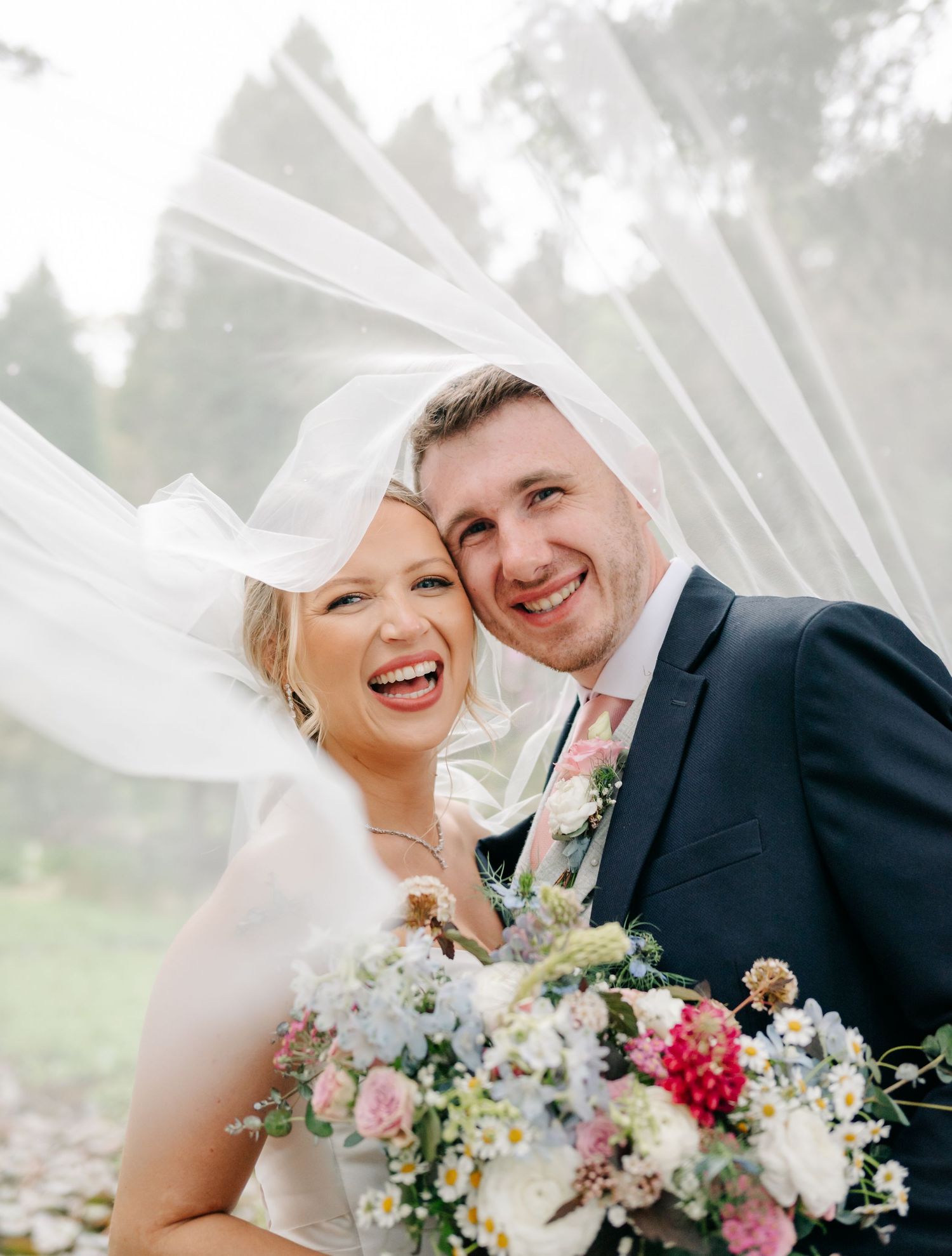 Happy bride and groom smiling under flowing veil, bride holding colorful spring bouquet on their wedding day.