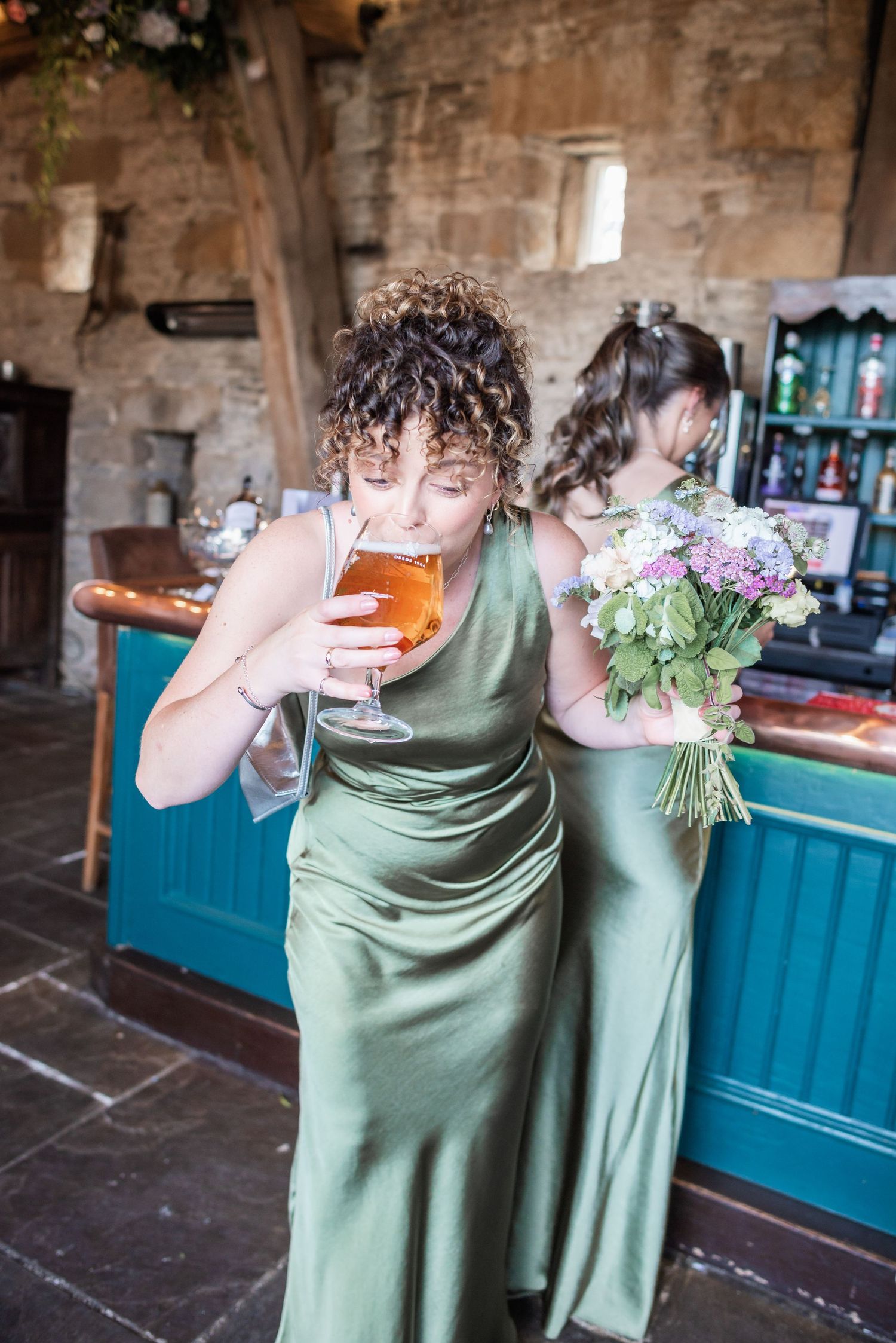 A person in a sage green formal dress takes a sip from a beer glass in a rustic stone interior.