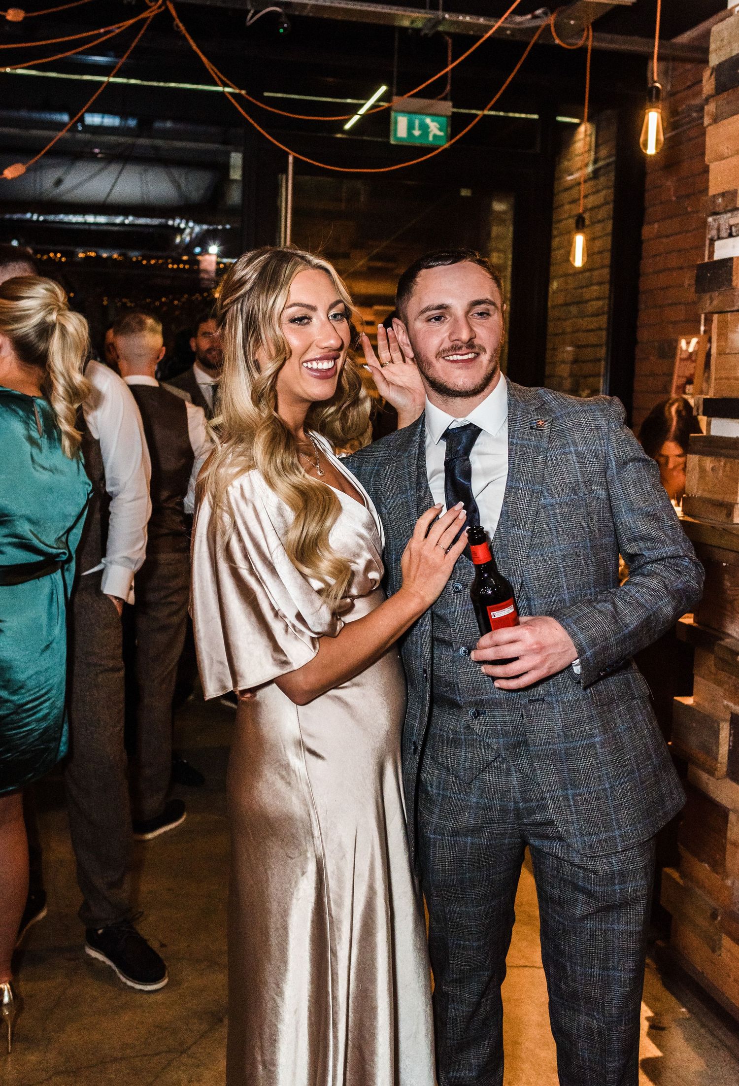 Couple in formal attire celebrating at an industrial-style venue with exposed brick walls and hanging lights.