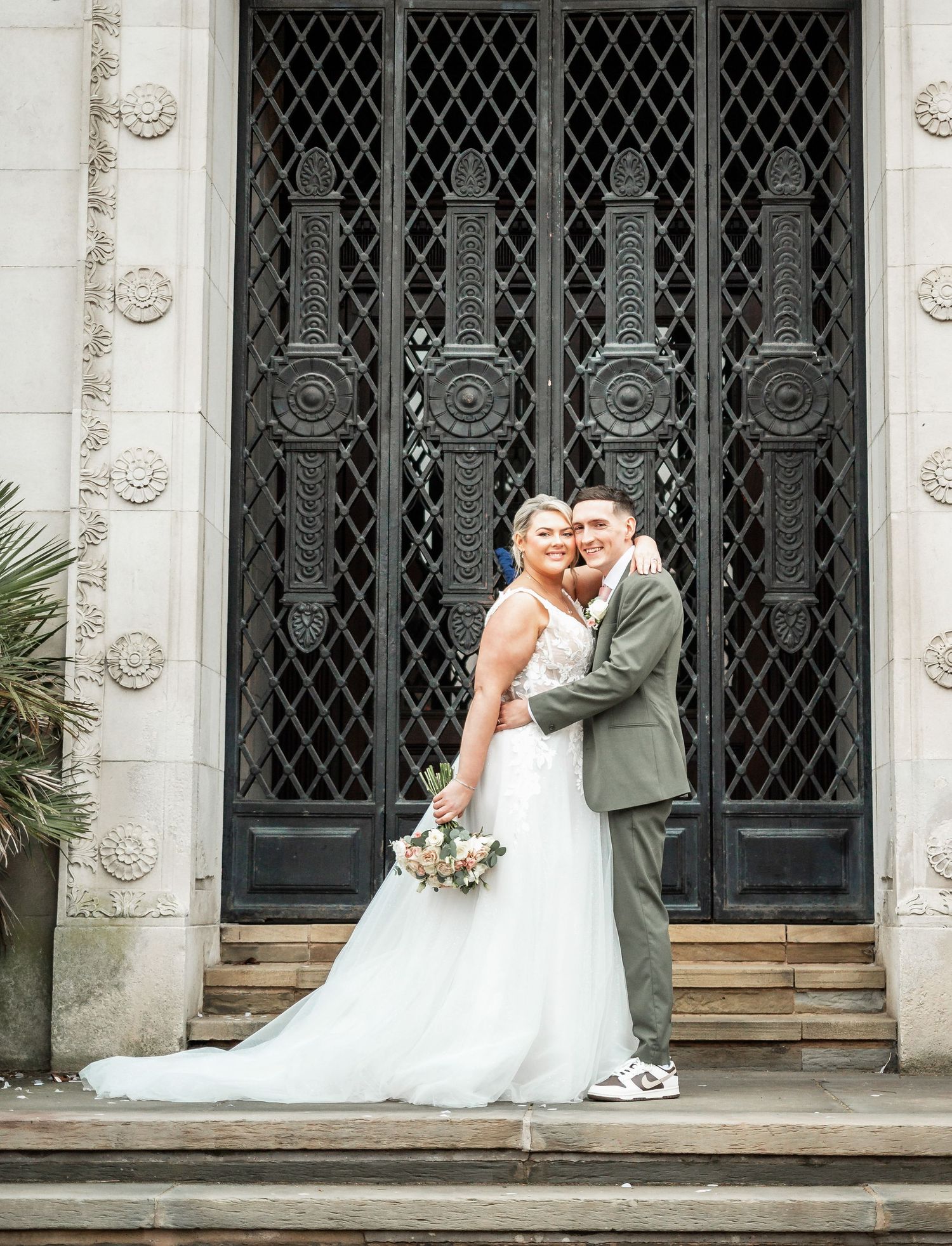 Wedding couple poses in front of ornate iron gate doors at historic building entrance.