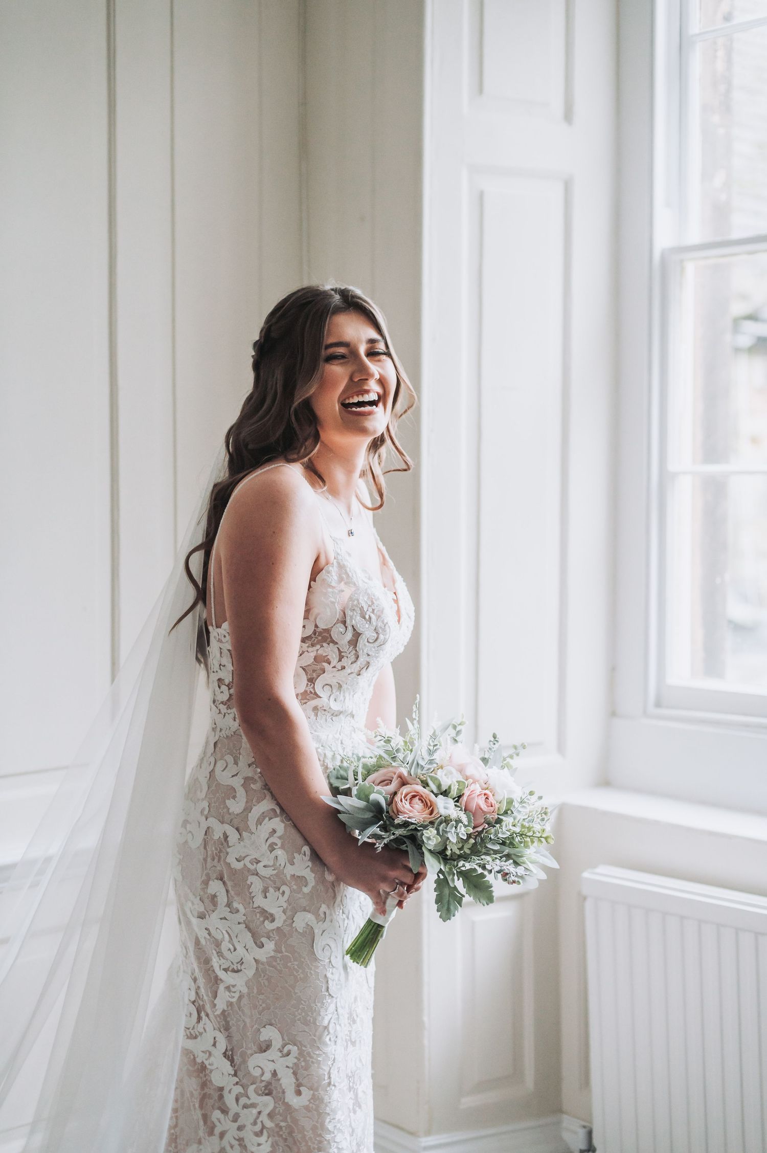 Bride in lace wedding dress laughs joyfully while holding a romantic blush and white bouquet by bright window.