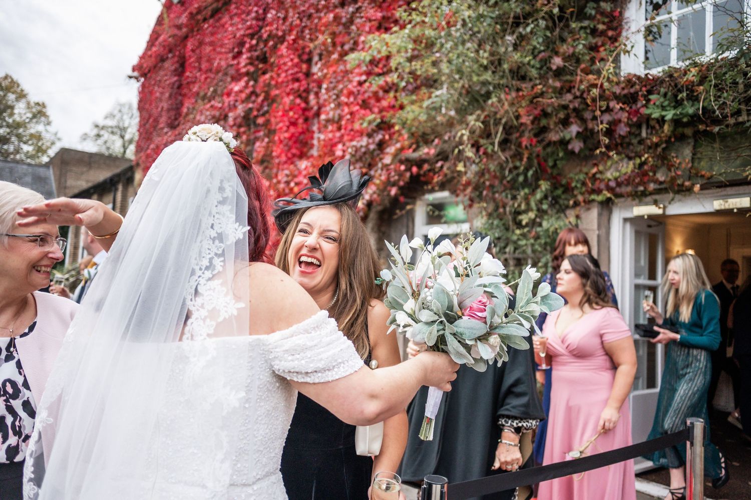 Wedding guests celebrating with joyful laughter outside a building covered in red fall leaves.