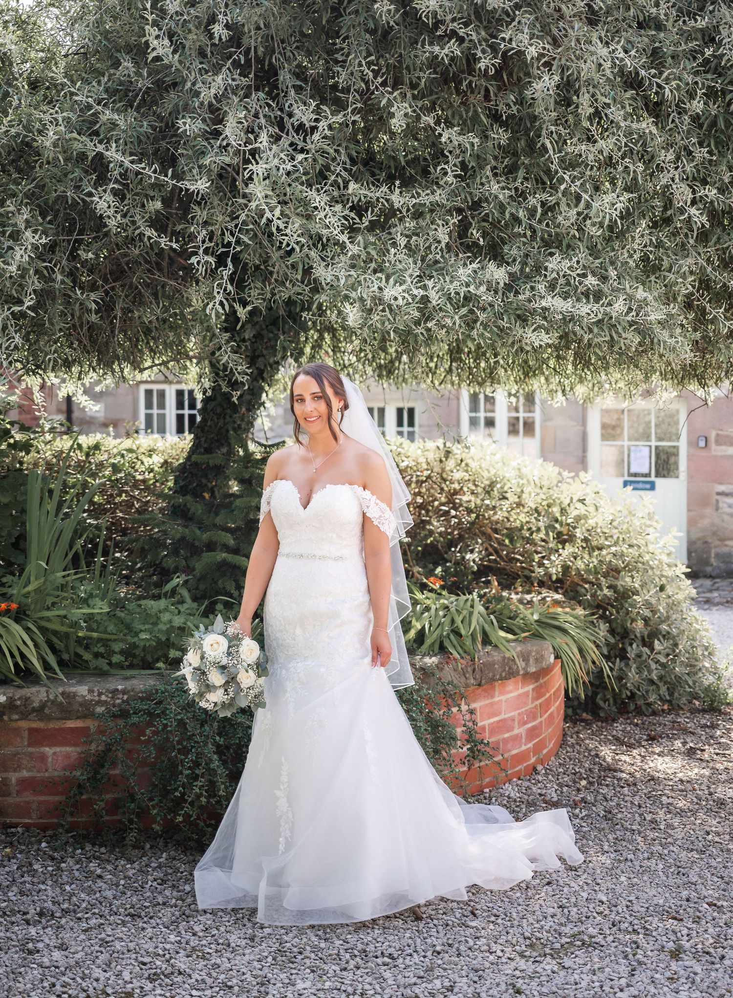 A bride in a fitted lace wedding dress poses in a garden setting with beautiful landscaping.