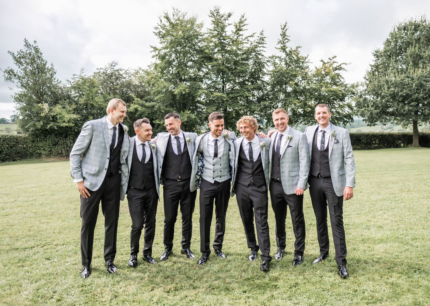 Groomsmen in coordinated gray suits and black ties standing on grass for formal photo.