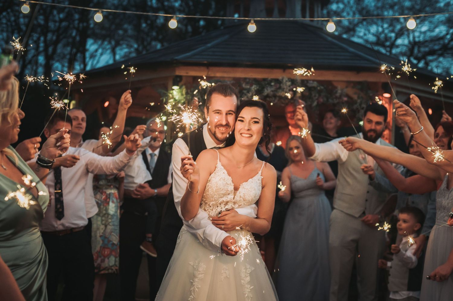 Newlywed couple and wedding guests dance beneath string lights at an outdoor evening reception celebration.