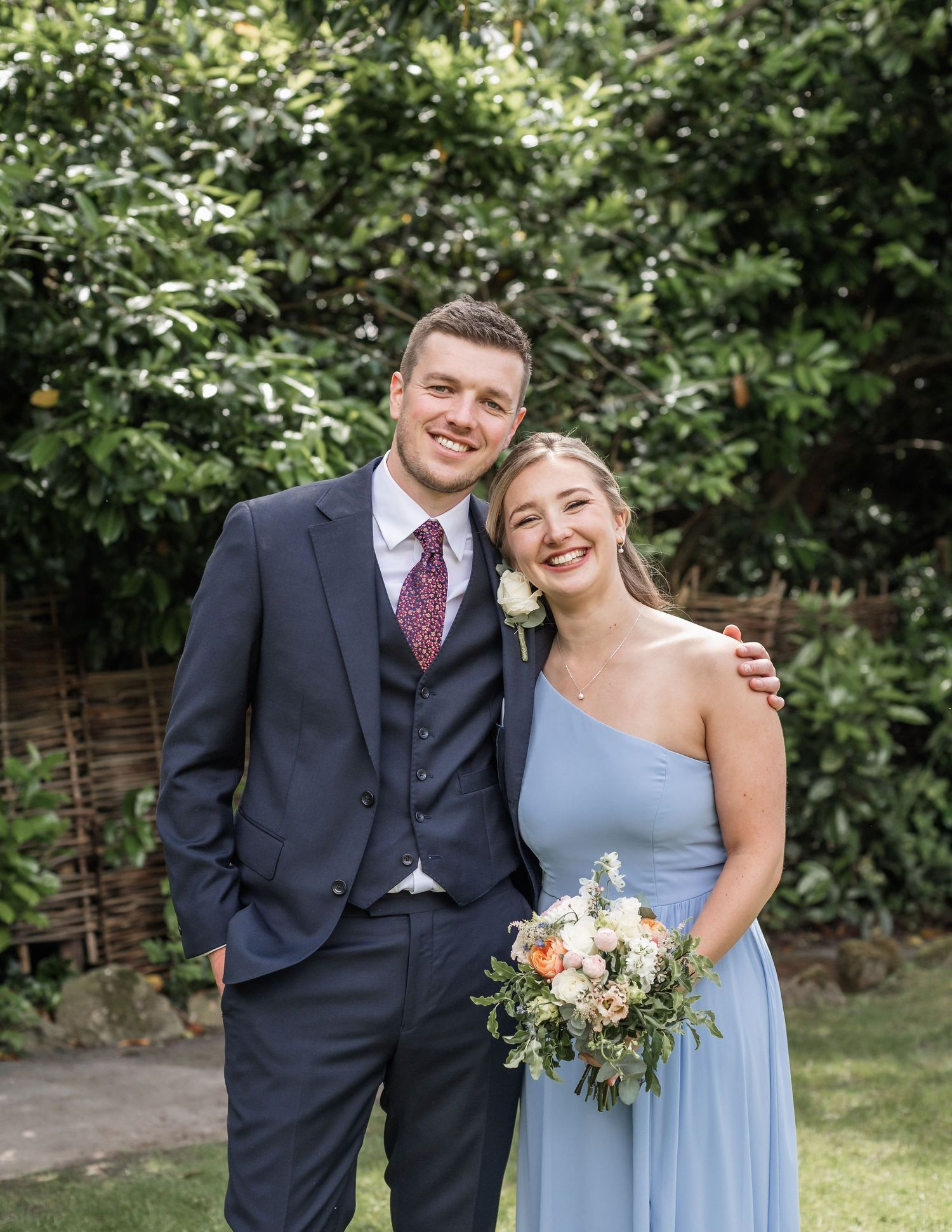 A couple in formal attire poses outdoors against a lush garden backdrop.