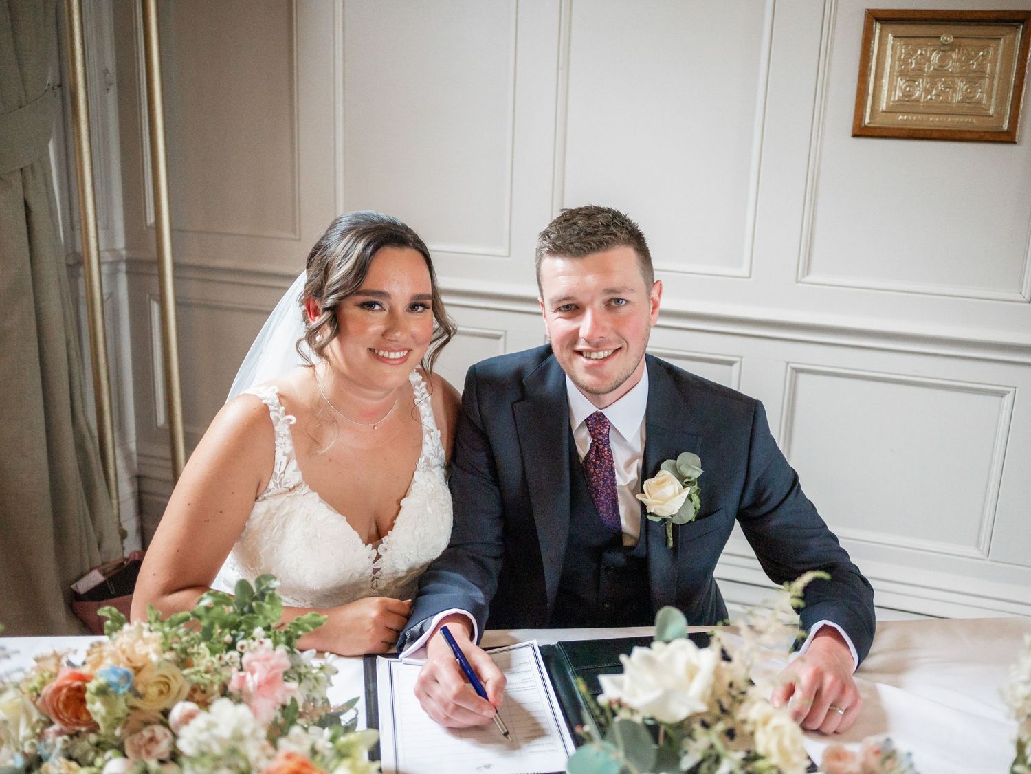 Newlyweds signing wedding documents together at an elegant indoor venue.