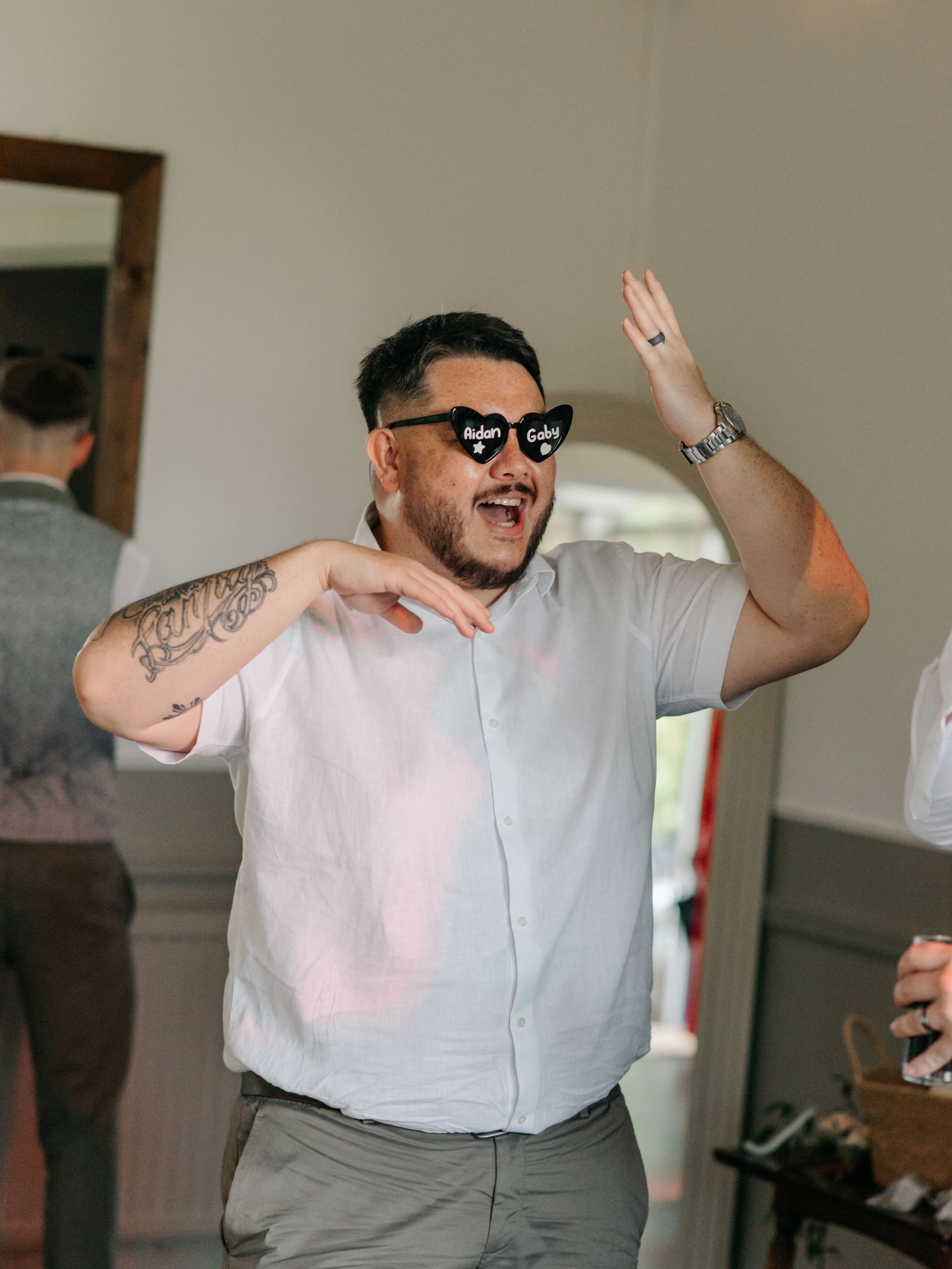 Wedding guest in sunglasses wearing grey shirt raises hand while dancing at reception celebration.