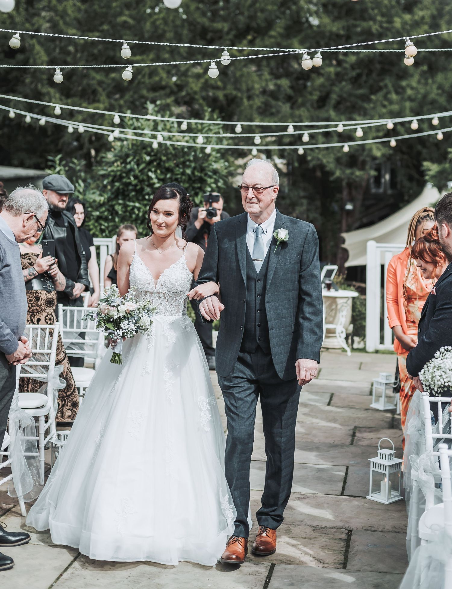Bride in white gown walks down aisle under string lights during outdoor ceremony.