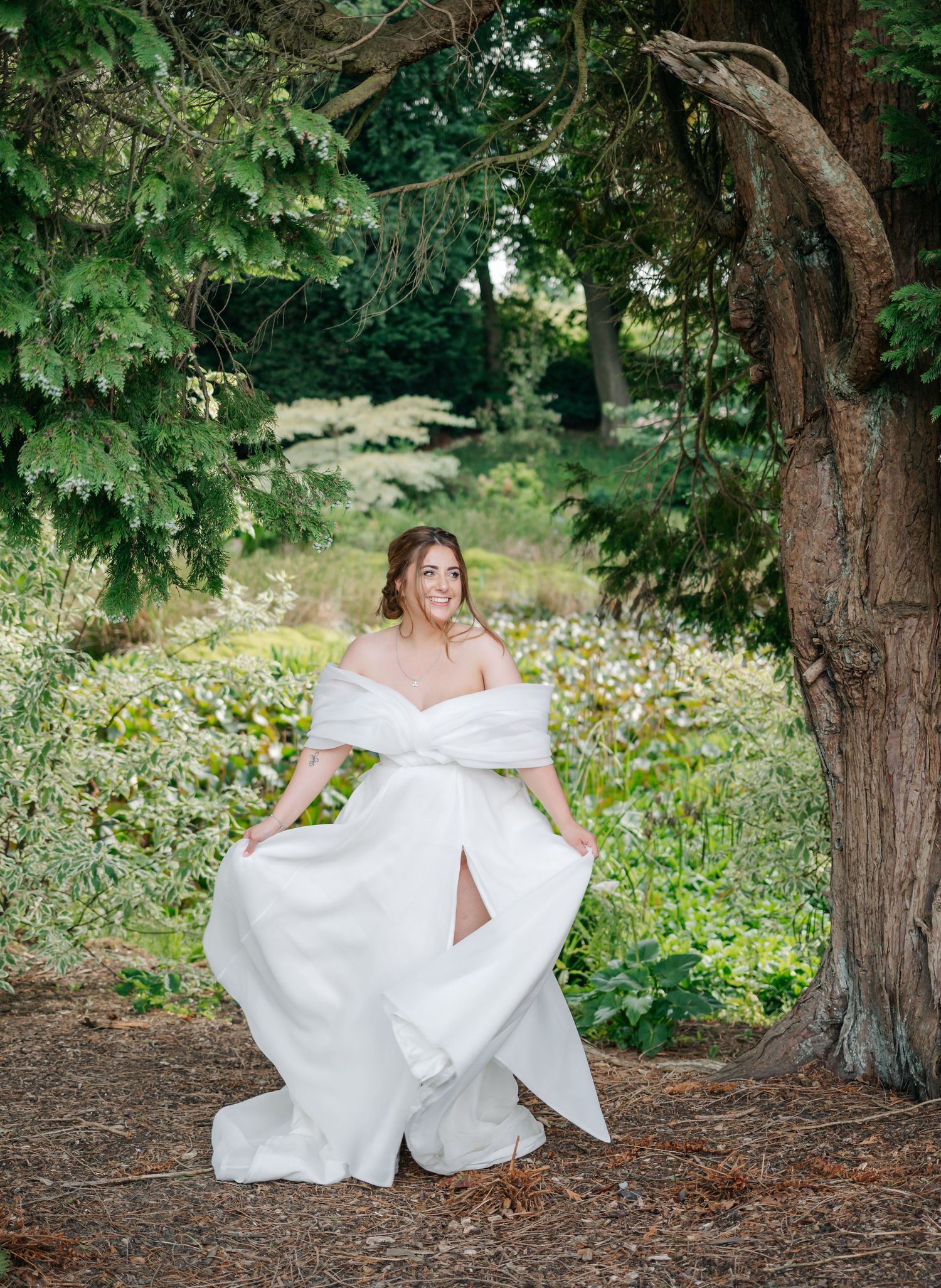 A bride in a flowing white off-shoulder gown poses gracefully among evergreen trees and wildflowers in a garden setting.