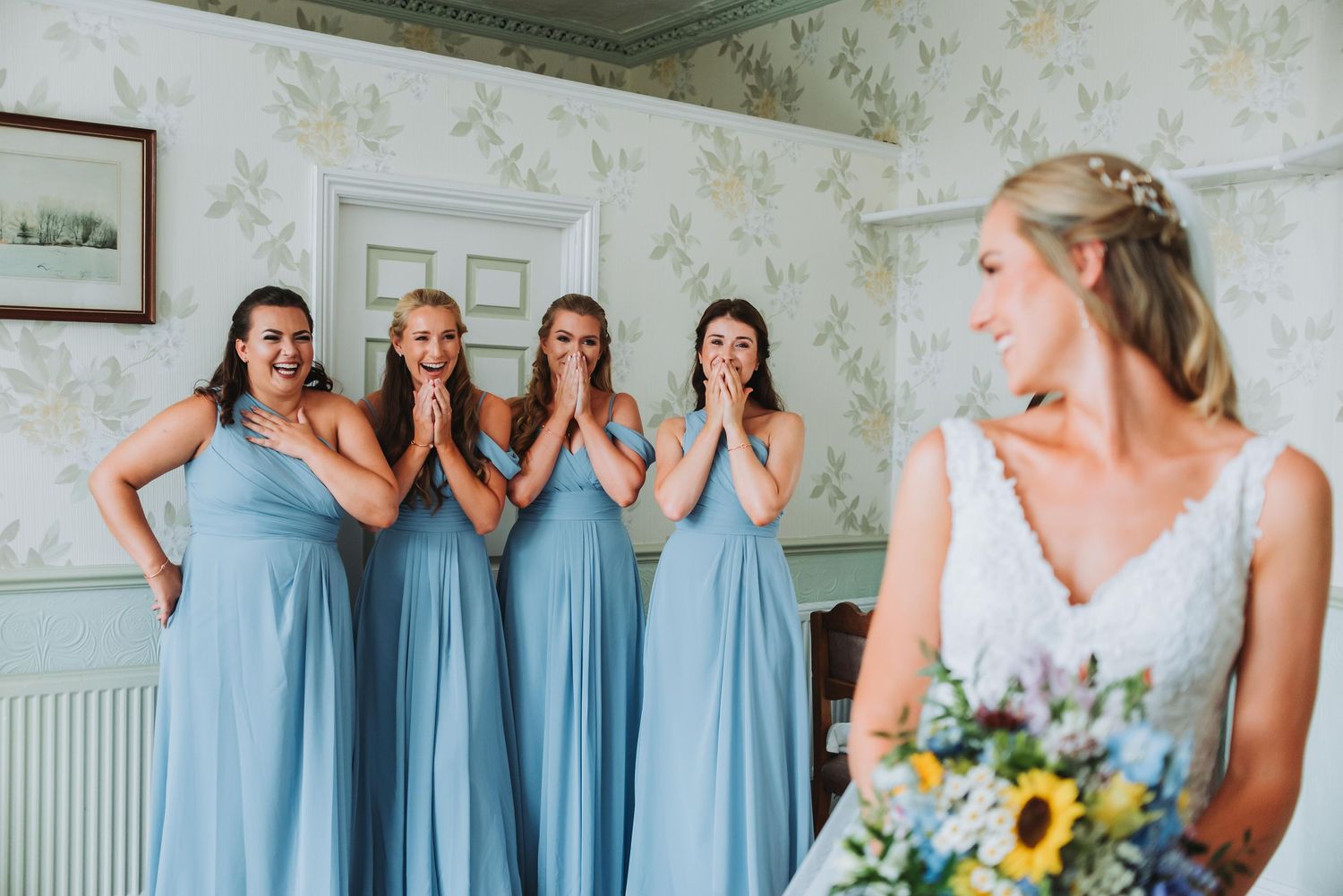 Bridesmaids in light blue dresses react with surprise to bride's reveal.