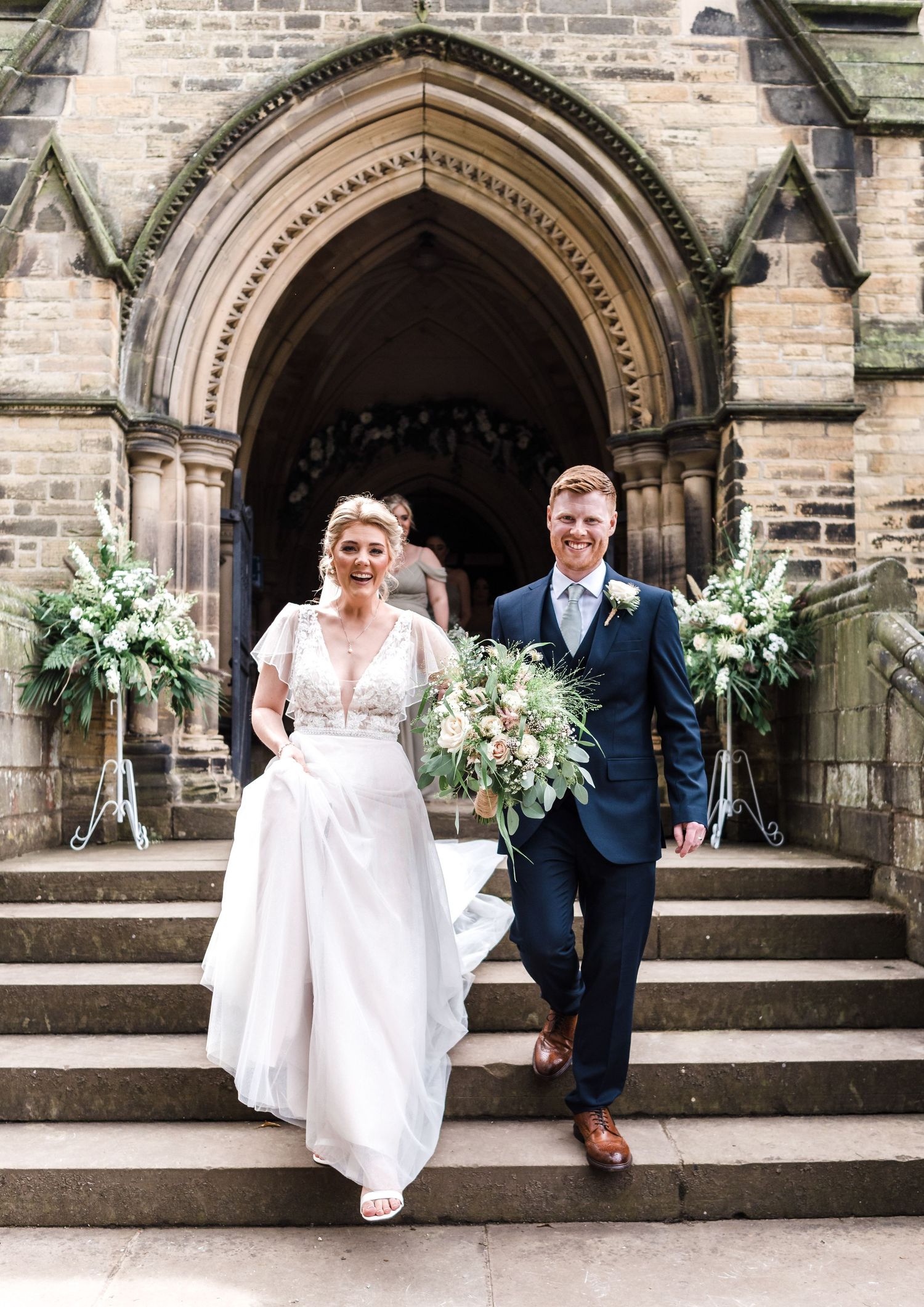 A couple descends stone church steps decorated with floral arrangements after their ceremony.