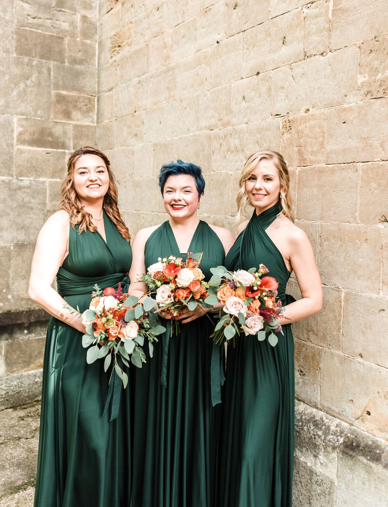 Three bridesmaids in emerald green dresses holding coral bouquets against stone wall.