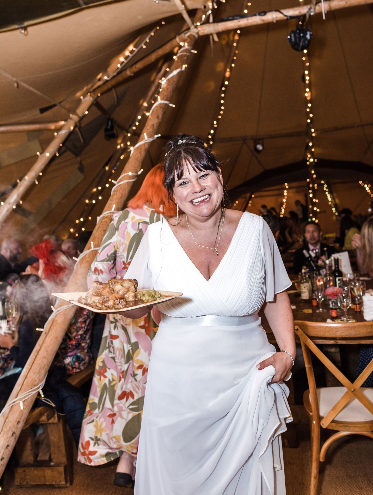 A joyful party guest in a white dress carrying a plate of food under string lights.