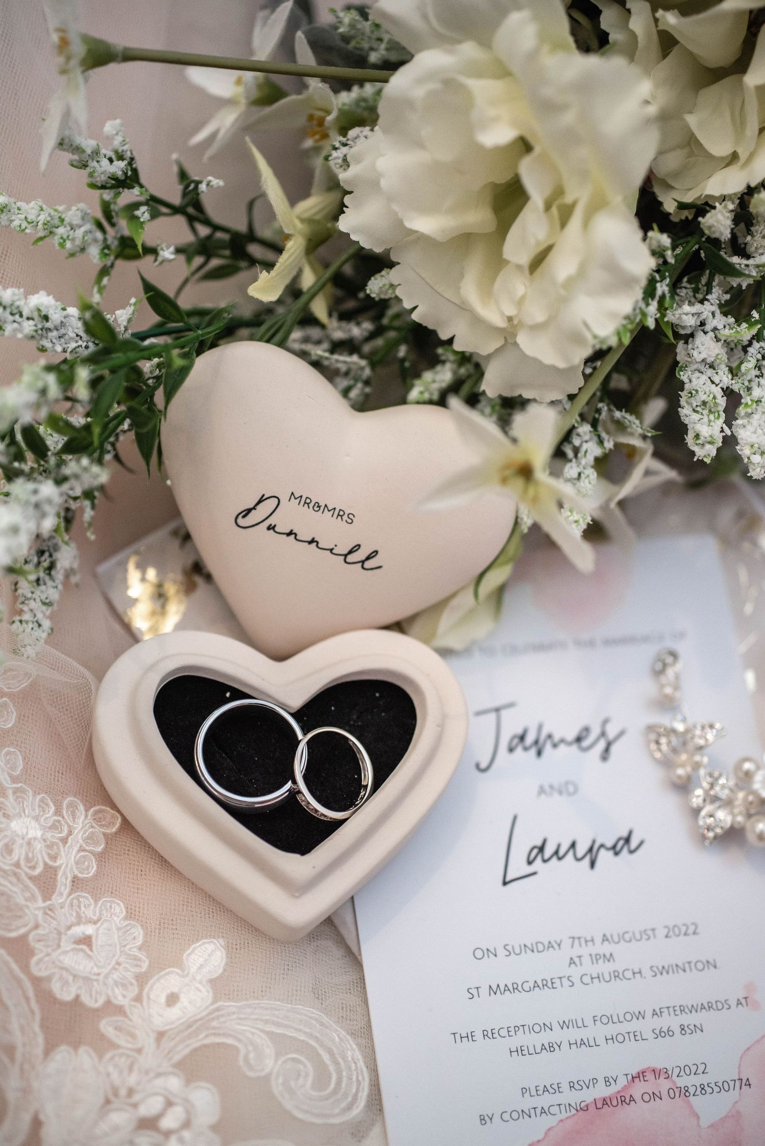Wedding rings in heart-shaped holder with white flowers and invitation details.