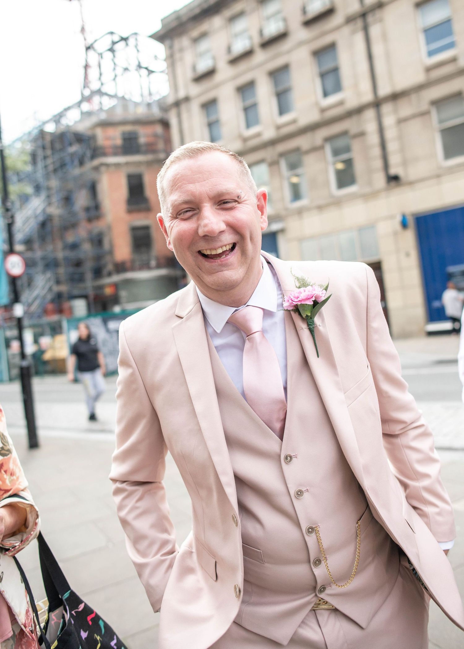 Person in a light pink suit and floral boutonniere smiling outdoors in an urban setting.