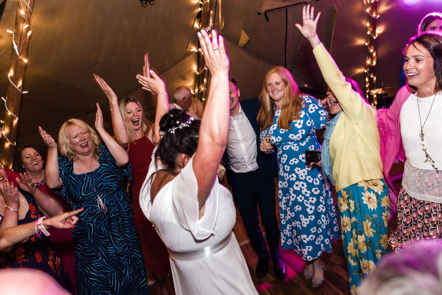 Wedding party guests dancing with raised arms under string lights.
