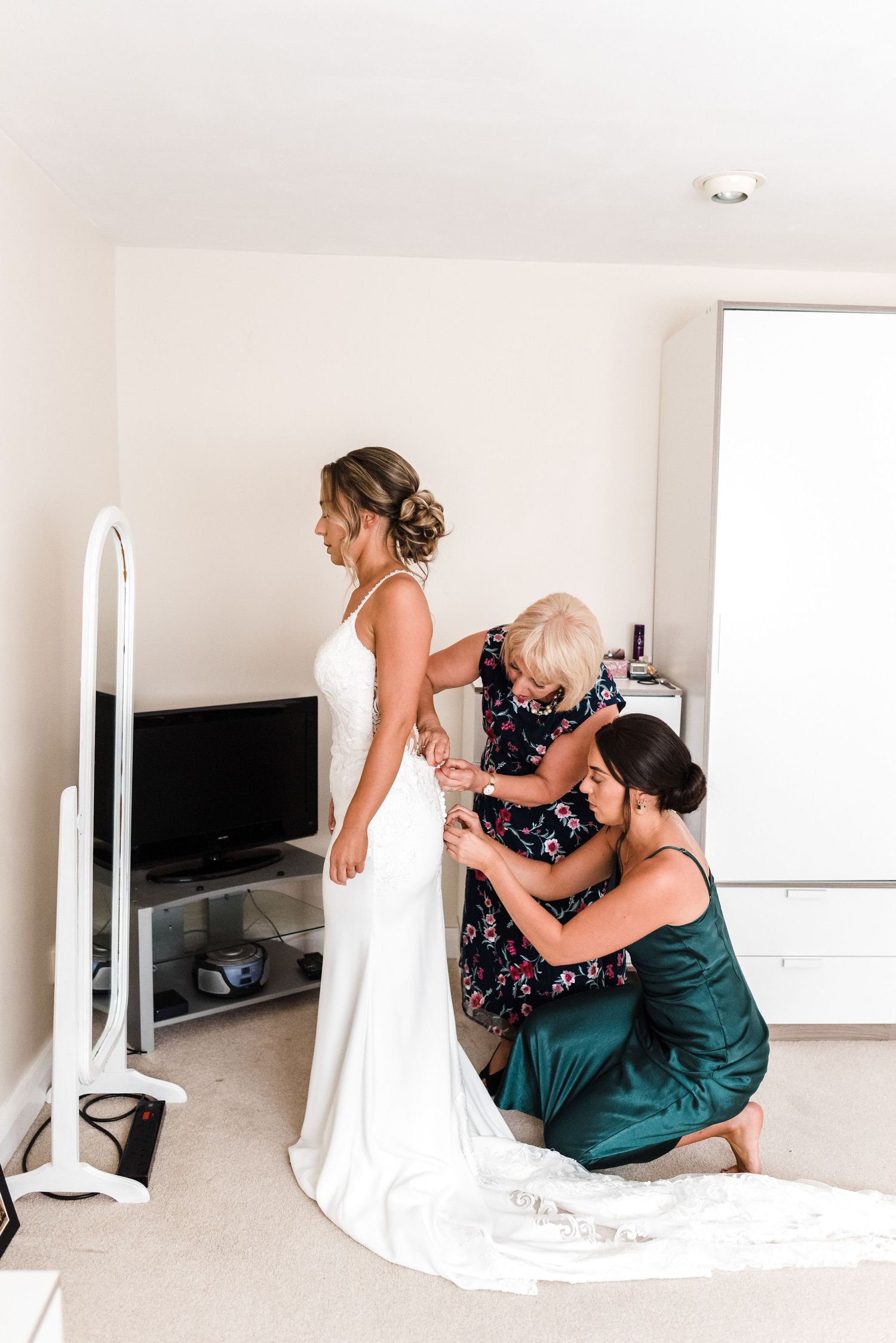 Helpers assist with adjusting a wedding dress during final preparations in a bright room.