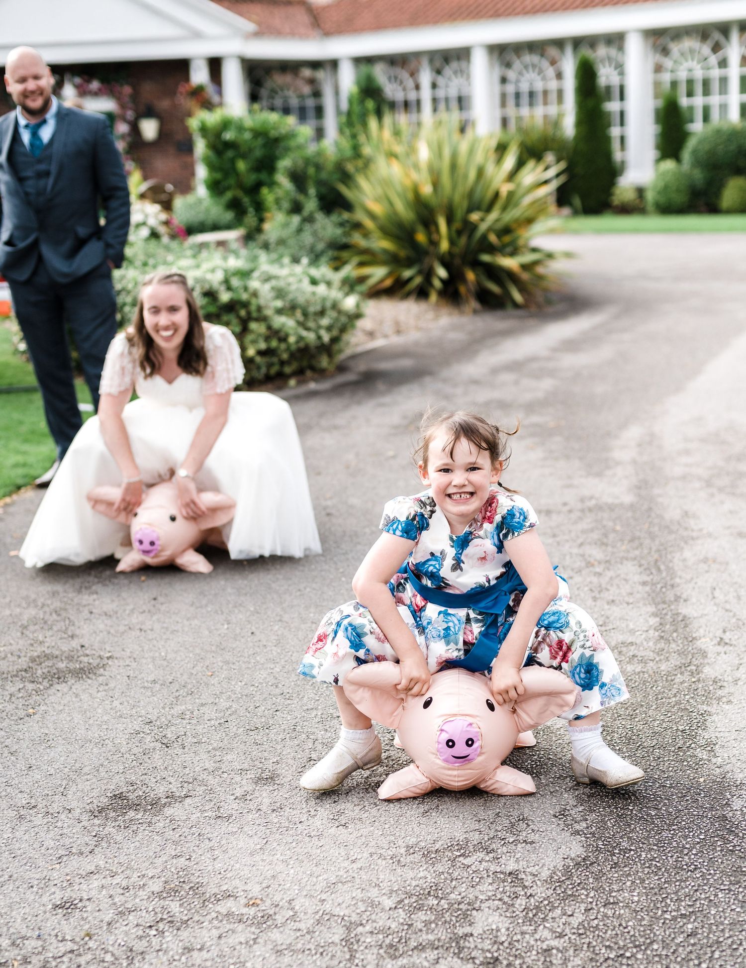Playful wedding photo with people sitting on stuffed pink pigs on a garden pathway.