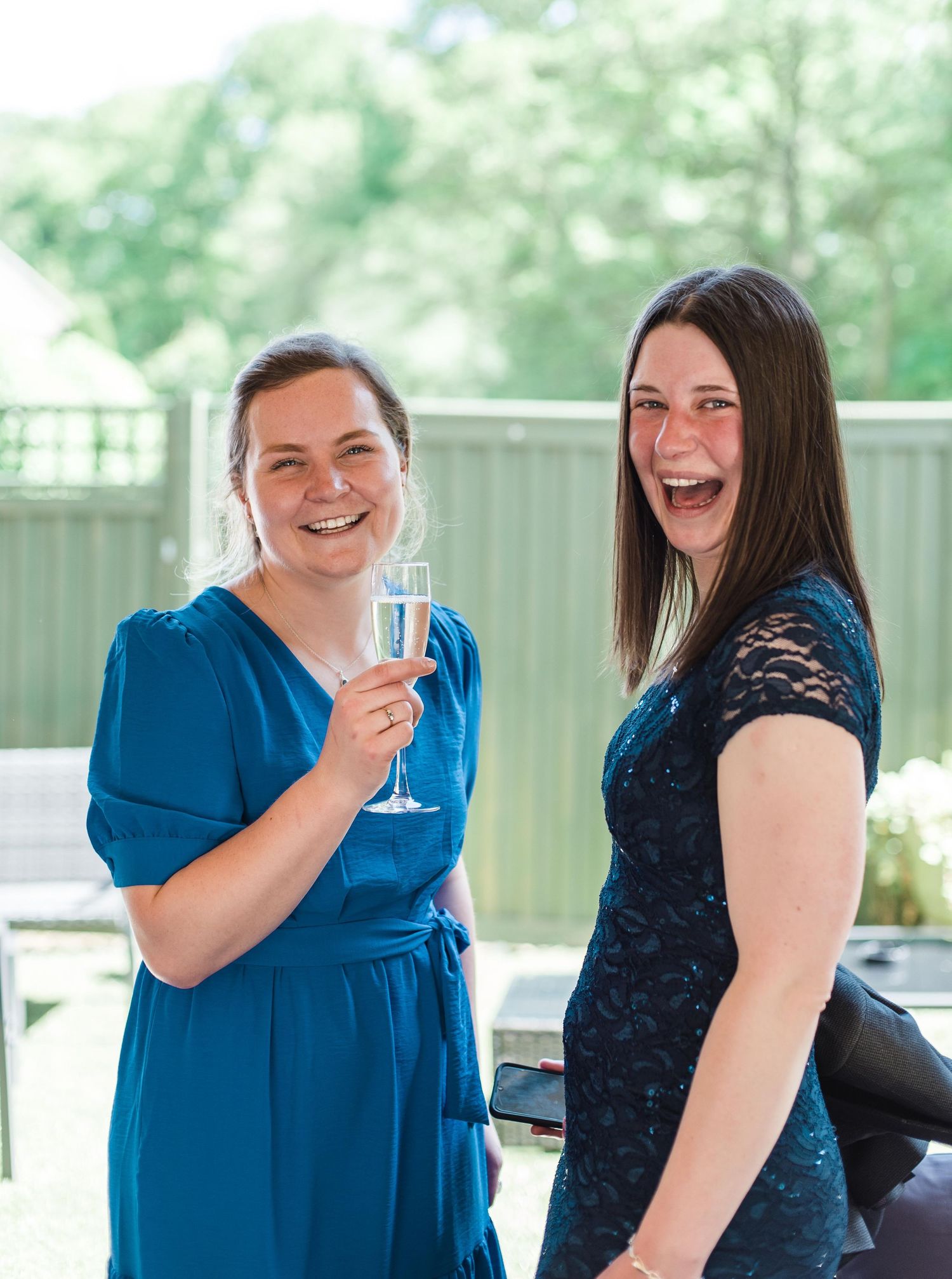 Two guests in blue and navy dresses sharing laughs at outdoor reception.