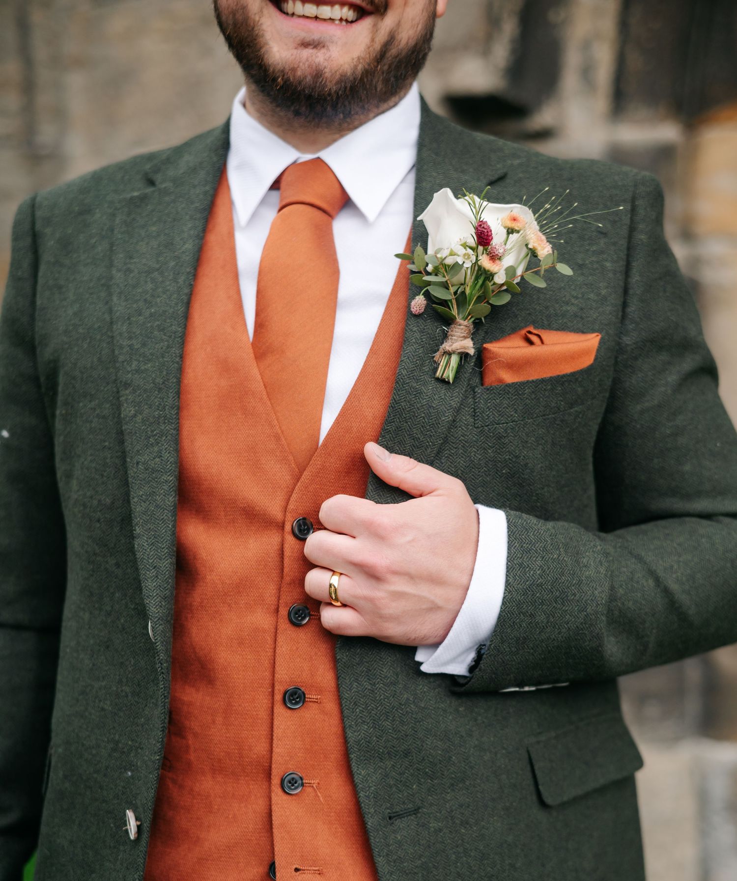 Close-up of a groom in a green tweed jacket, orange waistcoat and tie, with a floral buttonhole and pocket square.