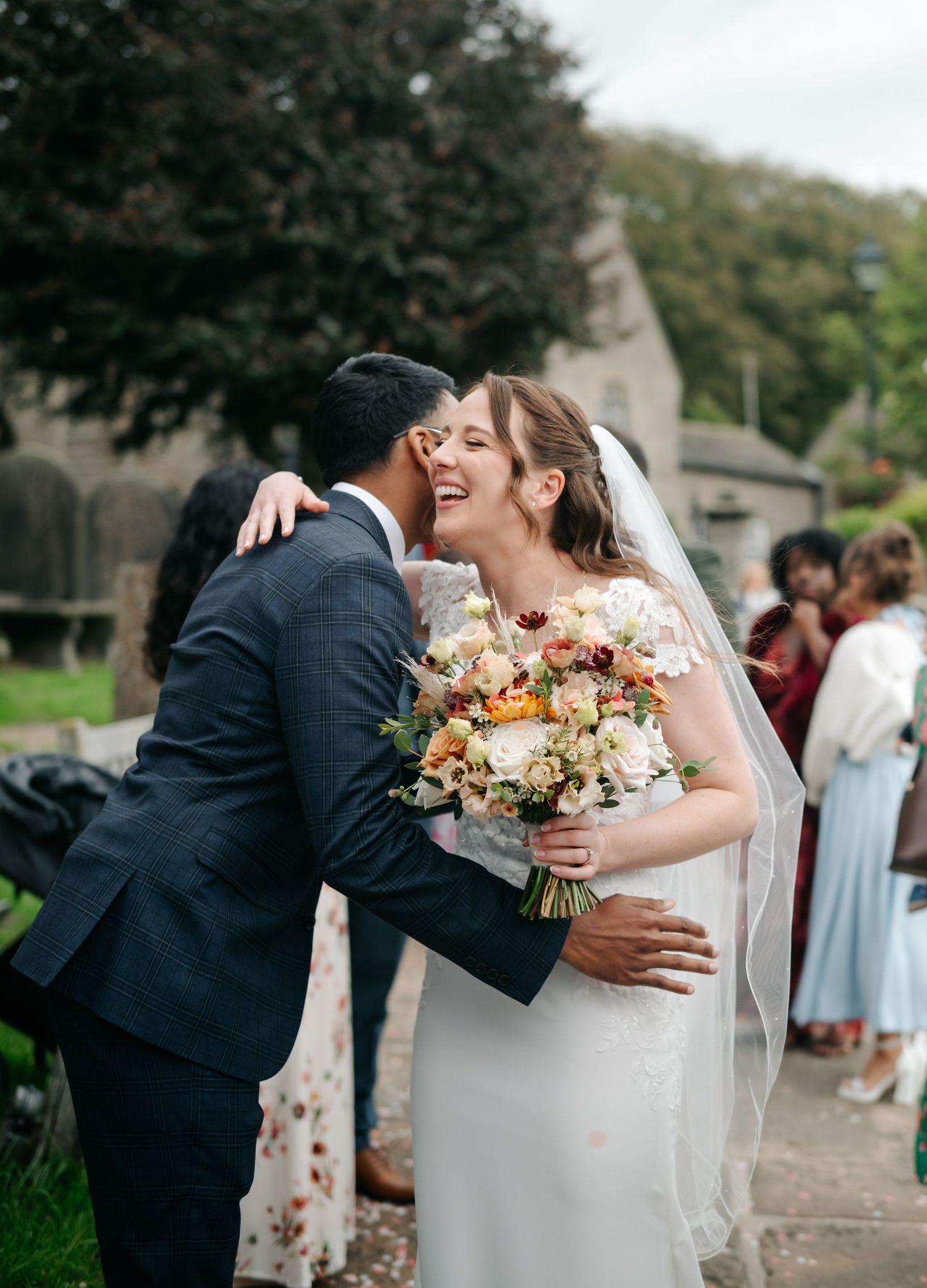 Smiling bride in a lace gown and veil holding a bouquet of peach and white flowers as she hugs a wedding guest in a navy suit.