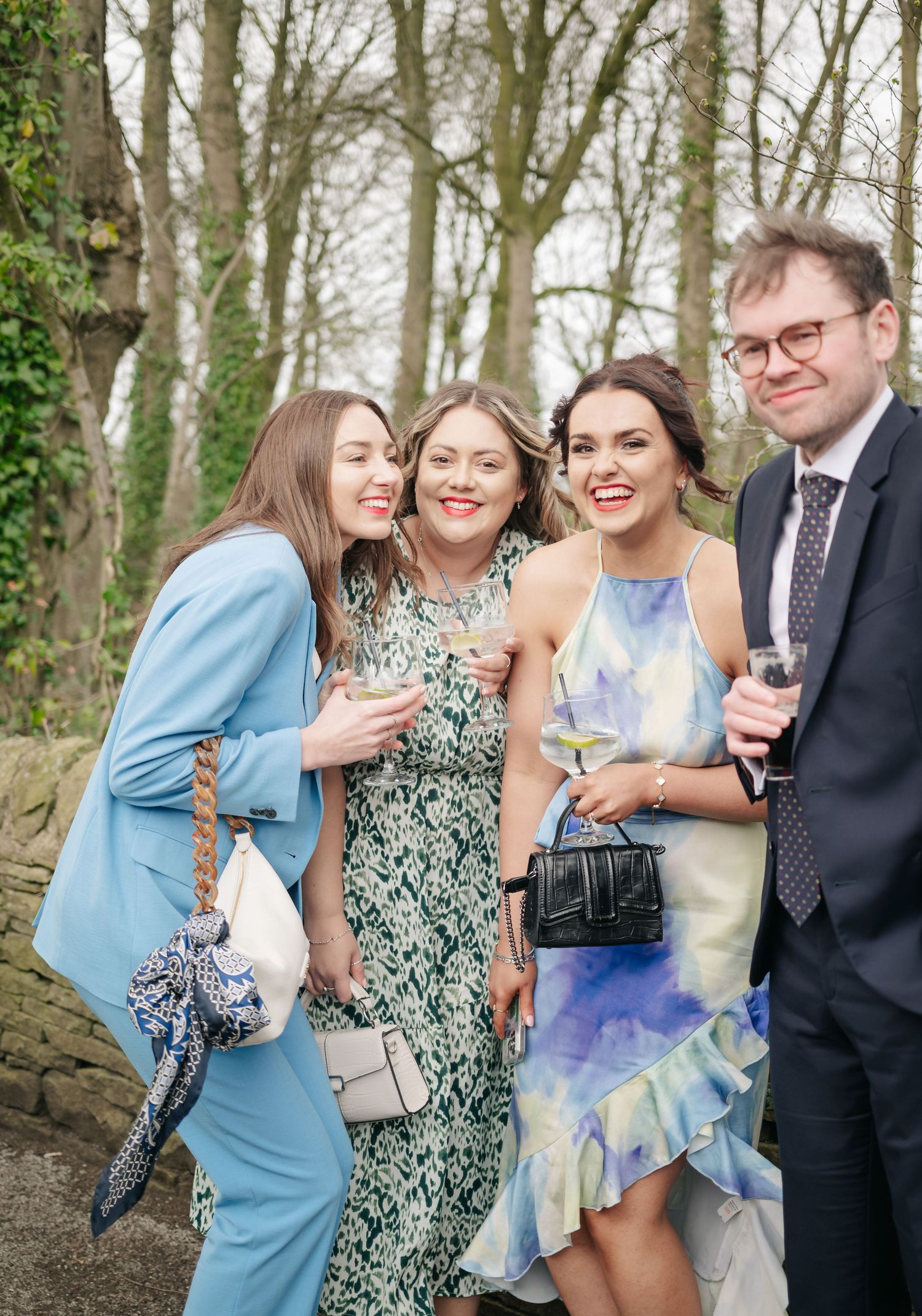 Wedding guests in floral dresses and formal attire celebrating outdoors among trees.