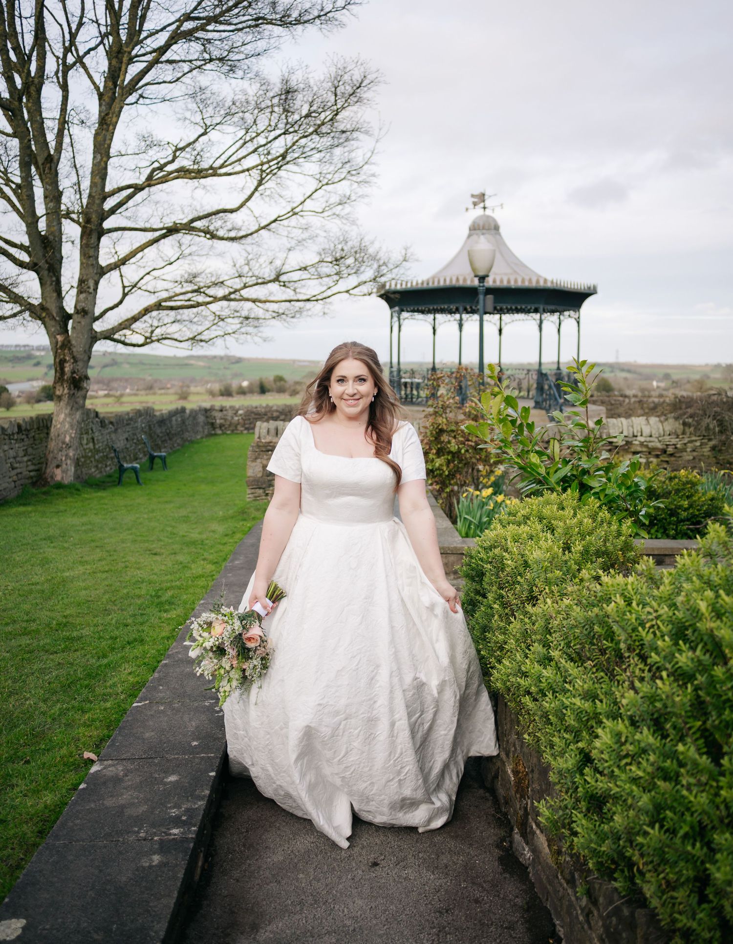 A bride in a white dress walks along a garden path with a Victorian gazebo in the background.