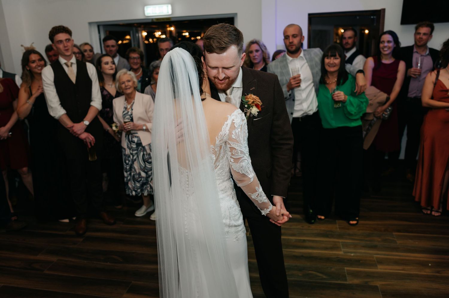 Wedding couple shares their first dance surrounded by guests in an indoor reception venue.