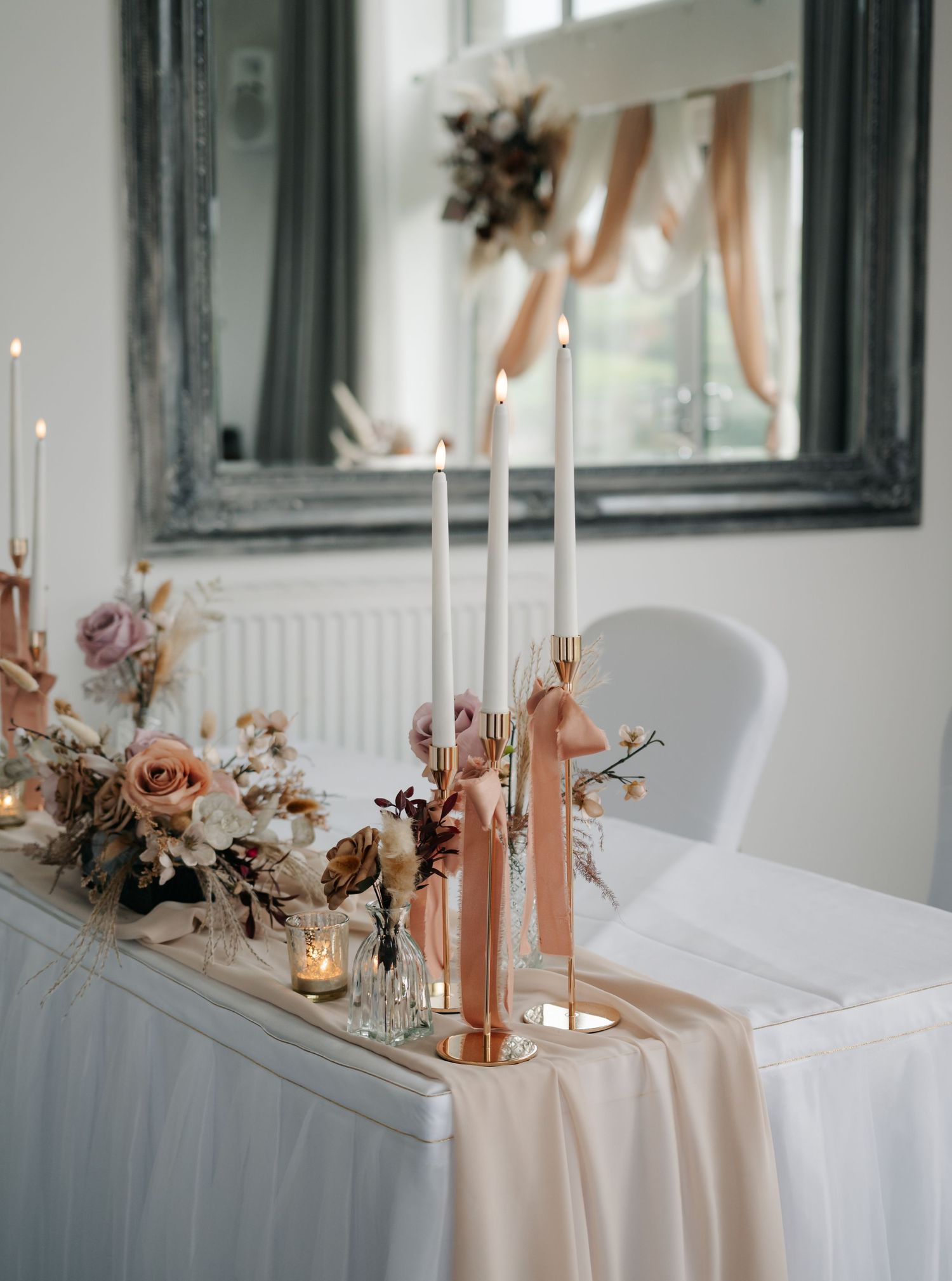 Elegant wedding table decor featuring peach candles, white linens, and dried floral arrangements in a bright venue.
