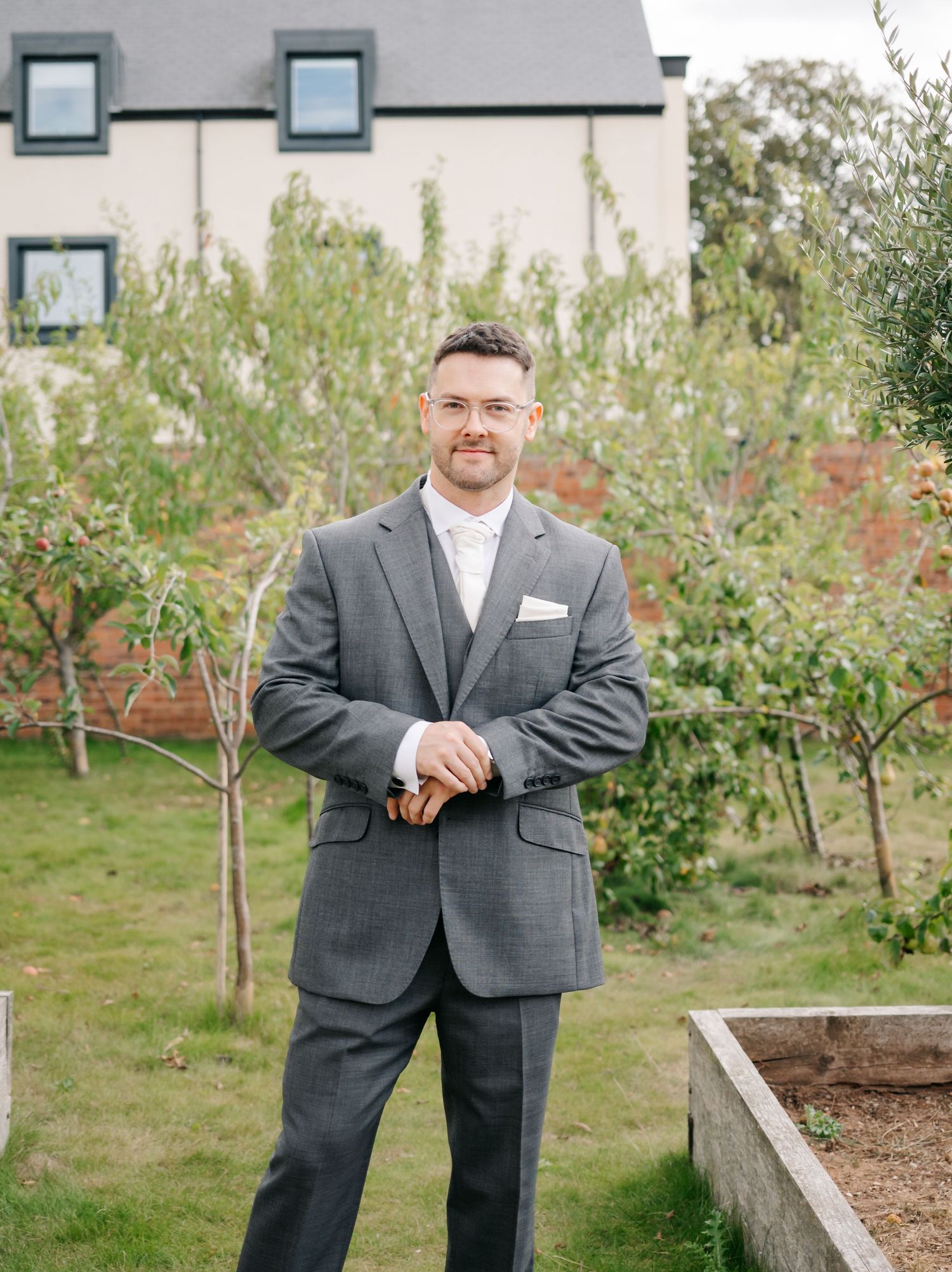 A man in a gray suit stands in a garden setting with a house in the background.