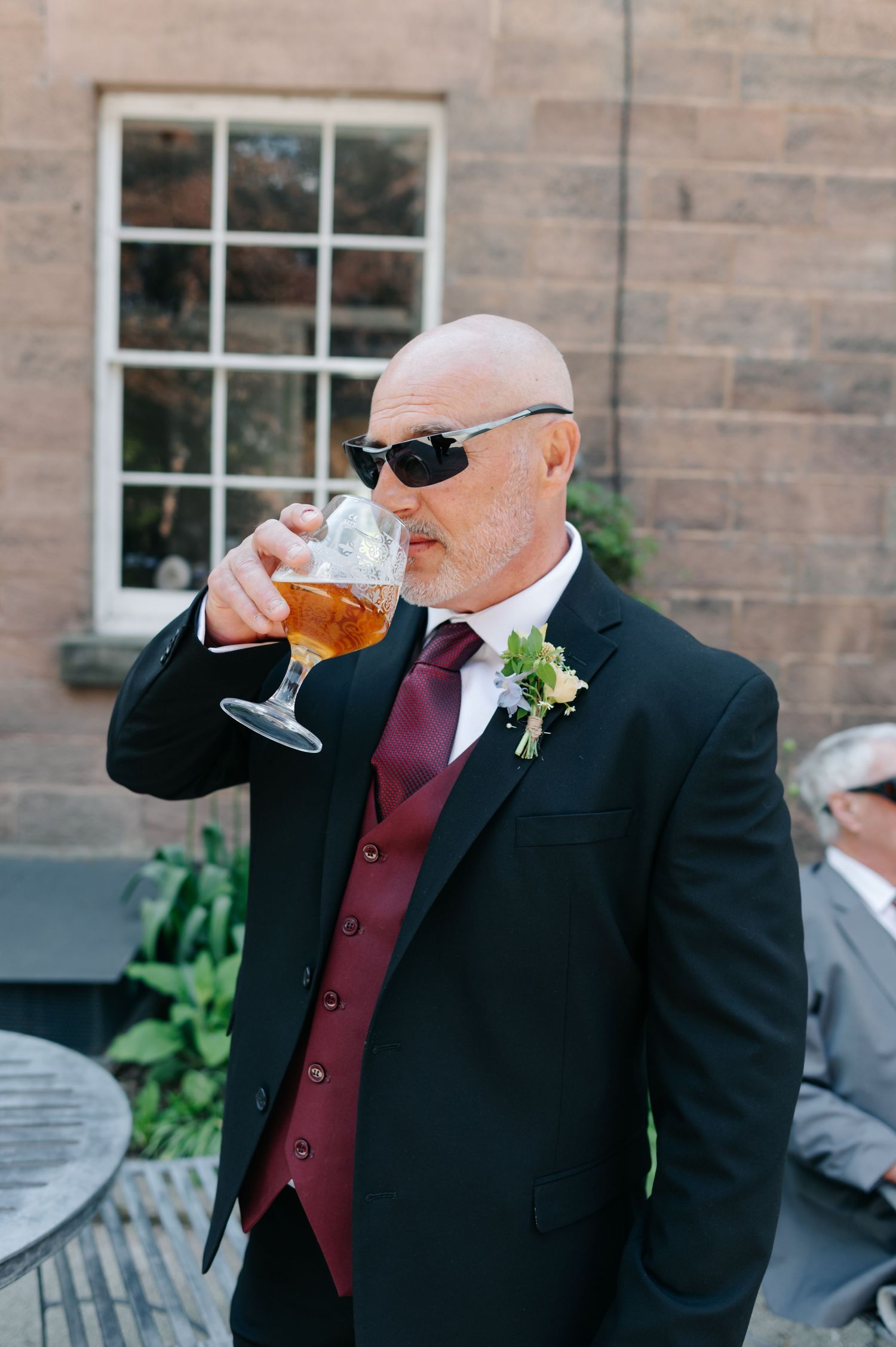 Wedding guest in black suit and sunglasses enjoying drinks on outdoor patio.